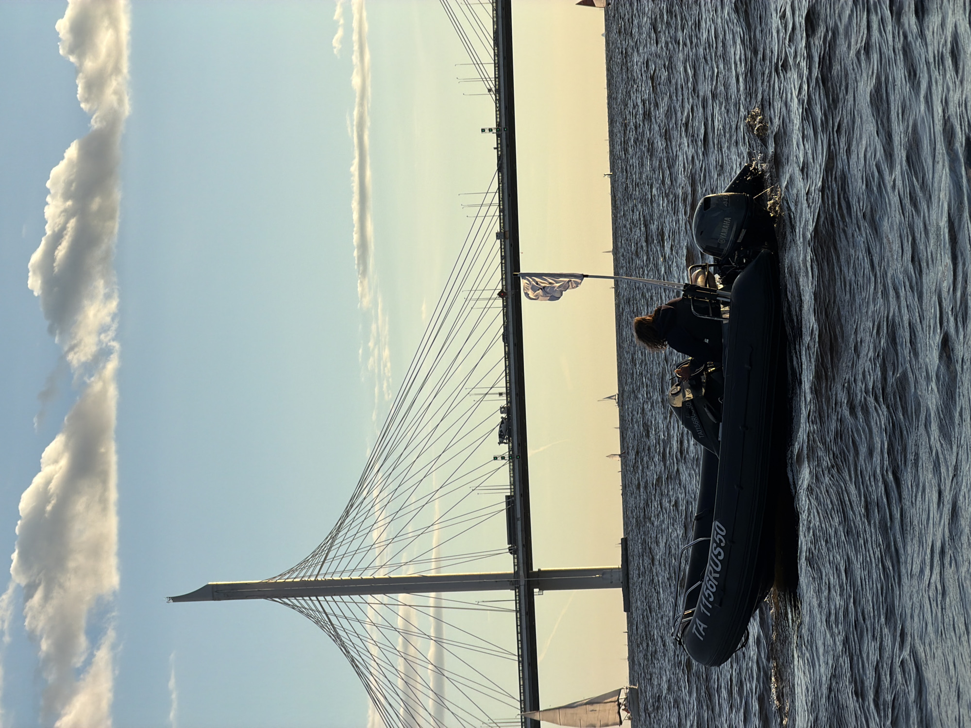 In Saint Petersburg, Russia, at sunset, a person is operating a dark-colored inflatable motorboat on choppy dark water. The boat has "TA 1156RUS50" written on its side. In the background, a large cable-stayed bridge spans the horizon, with vehicles visible on its roadway. Several sailboats are scattered across the water in the distance, and a white flag with a crest is visible on a pole in the middle of the boat. The sky is a light blue with scattered white and gray clouds.