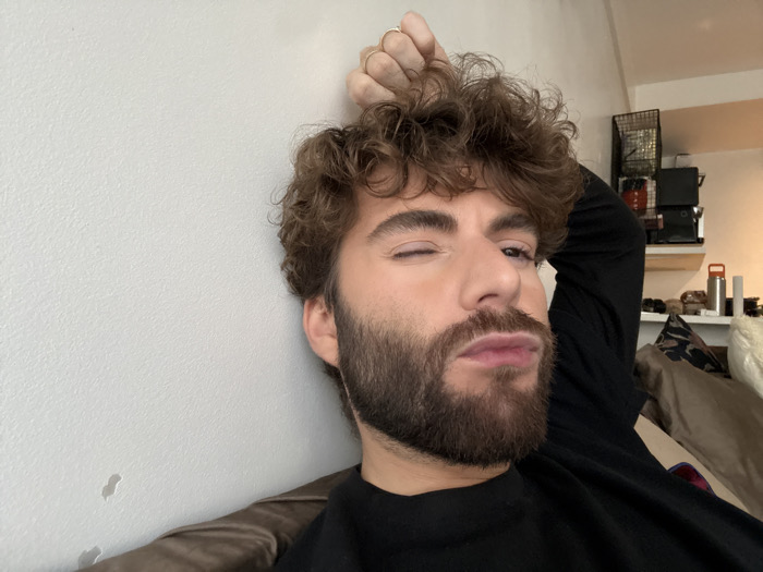 The scene depicts an adult male subject indoors, within a residential environment in Paris, France. The male has light skin, brown curly hair, and a full, dark beard, and is wearing a black long-sleeved garment, possibly a turtleneck. He is seen reclining, winking his right eye, and pursing his lips. His right hand, featuring a ring on the fourth finger, is raised and positioned near the top of his head. The immediate background to the left is a plain, light-colored wall with minor textural variations and faint marks. To the subject's right and rear, a brown upholstered surface is visible, alongside an interior backdrop containing a dark metal wire shelving unit and a lighter-colored wall shelf. These shelves hold various indistinct household items, including what appears to be a white and orange water bottle, and other dark-toned objects. A patterned cushion is partially visible on the upholstered surface.