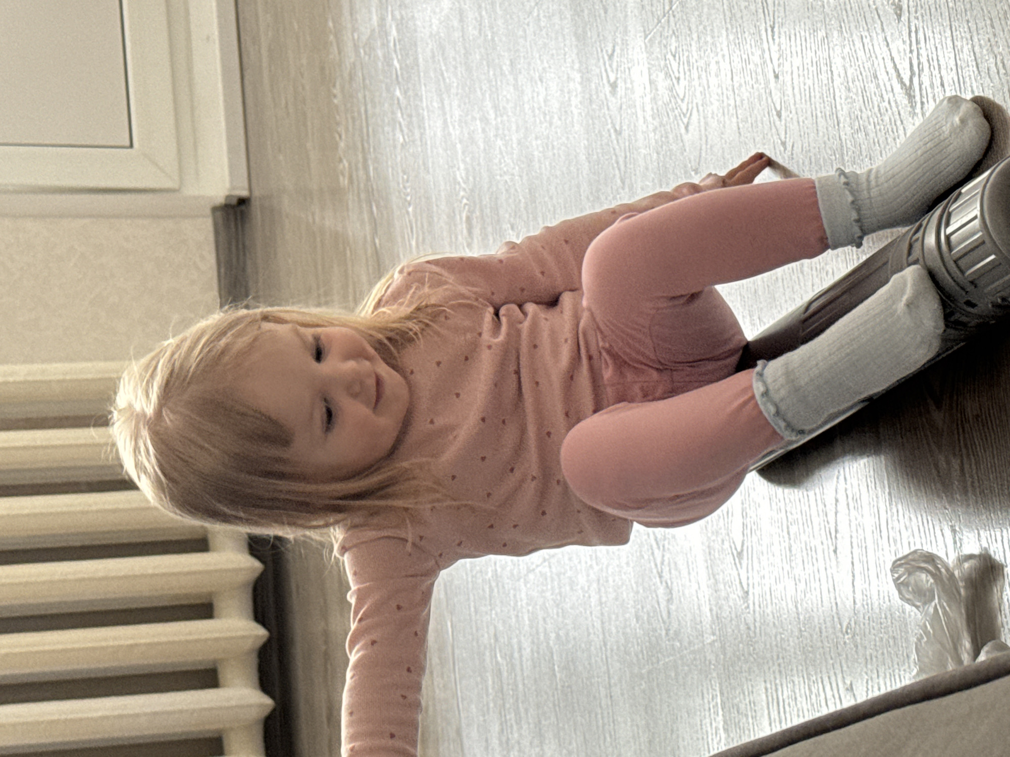 A young child with blonde hair lies on a wooden floor. She wears a pink long-sleeved shirt with a small heart pattern and matching pink leggings, along with light blue socks. Her feet rest on a dark gray cylindrical object with ridges. A radiator is visible to the left, and part of a window frame is above it. A crumpled transparent plastic bag sits near the bottom right corner of the image. The location and specific environment within the city of Noviy, Russia, cannot be determined.