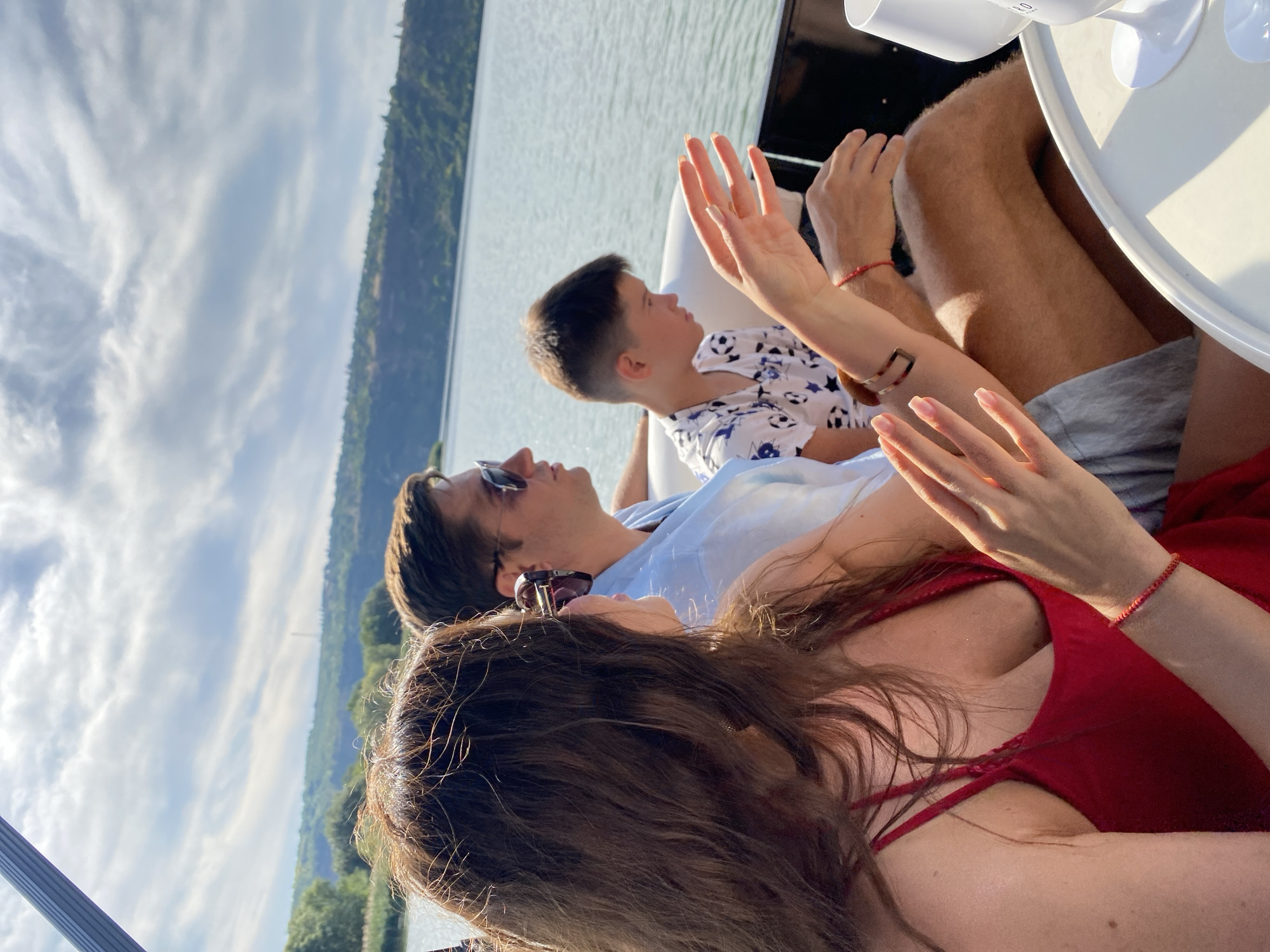 In Molovata Nouă, Moldova, three individuals are seated on a boat. On the left, a woman with long brown hair and sunglasses, wearing a red dress, is gesturing with her hands. Next to her, a man in a light blue shirt and sunglasses is looking forward. To the right of the man, a young boy wearing a white t-shirt with a black pattern is also looking ahead. The boat is on a body of water with a hilly, tree-covered landscape in the background under a cloudy sky.