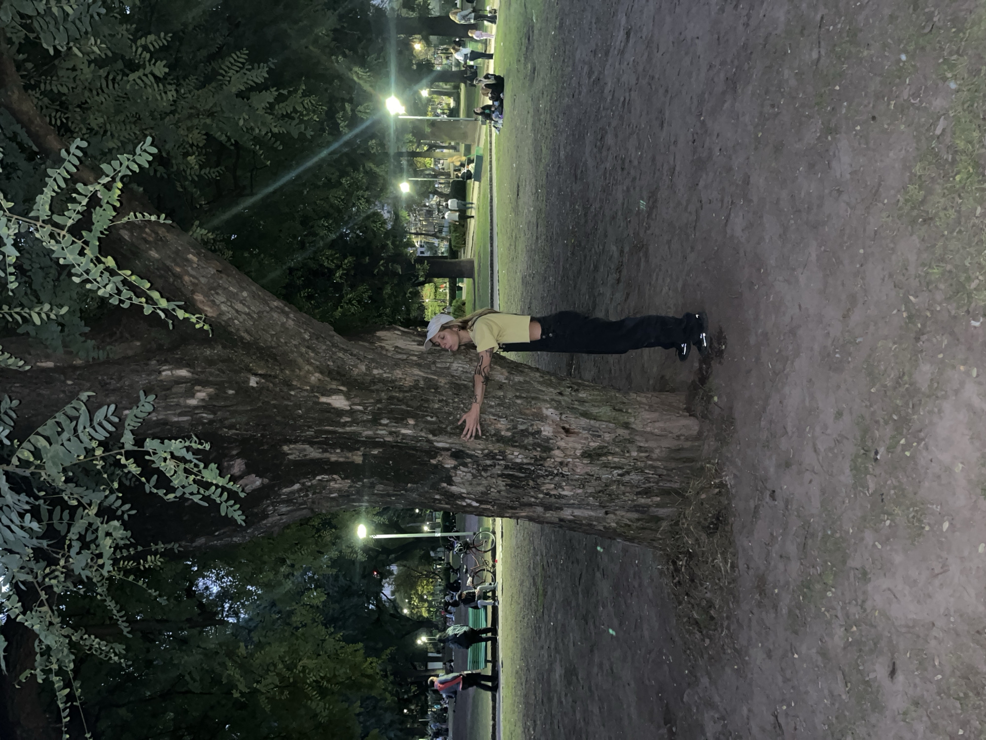 The image, taken in what appears to be a park in Buenos Aires, Argentina, shows a person embracing a large tree. The person, wearing a light-colored cap, a yellow crop top, and dark pants, is standing next to the tree trunk with their arms wrapped around it. The tree is situated on the edge of a park pathway. In the background, a park environment is visible with numerous people. There are streetlights illuminating the area. The ground is primarily dirt and grass. The photo is taken at night.