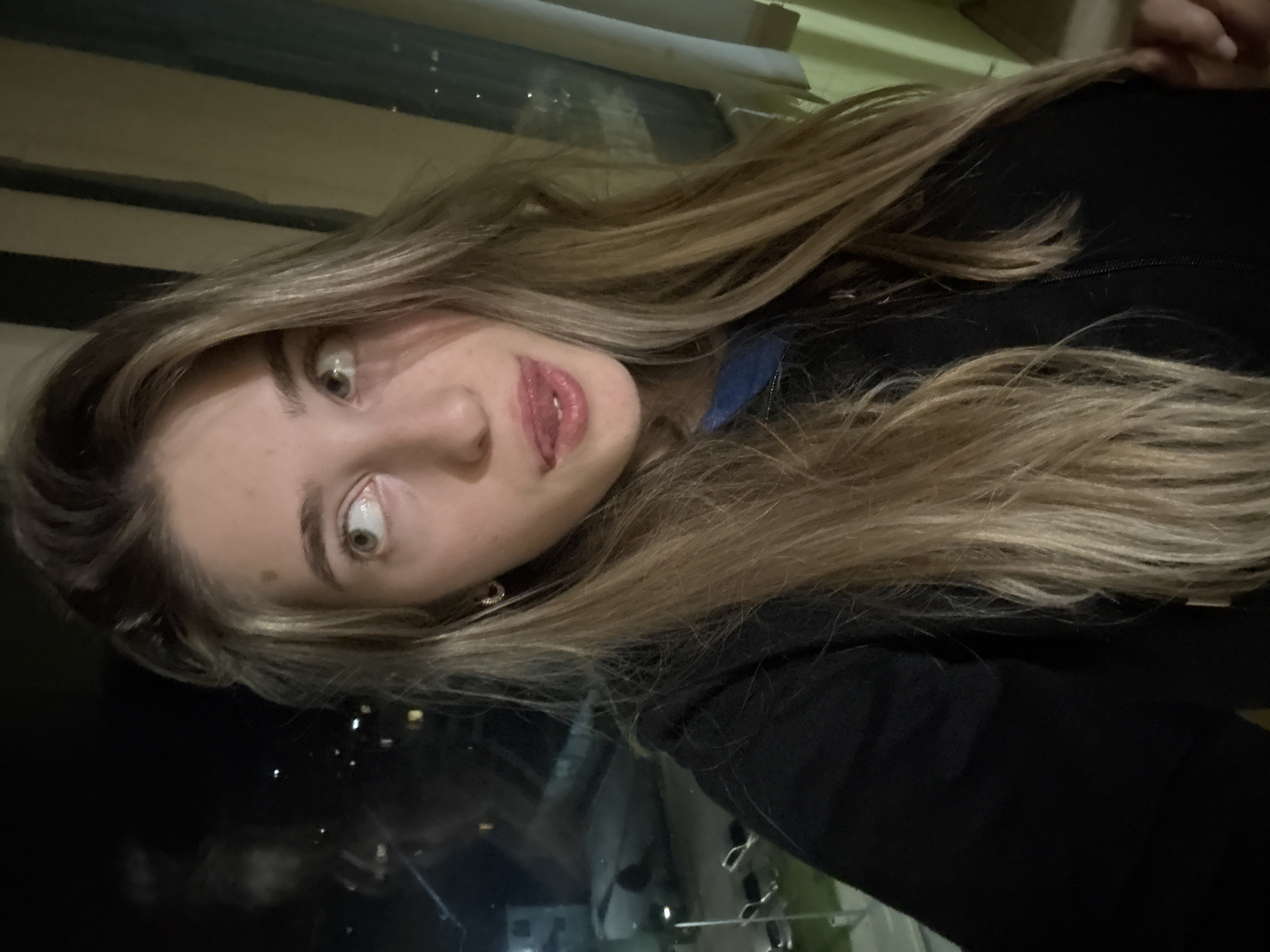 A woman with blonde hair is looking at the camera with wide eyes and a slightly open mouth. She is wearing a black jacket and has an earring in her left ear. The background shows a dark window with reflections of lights from the city of Unknown, Unknown.