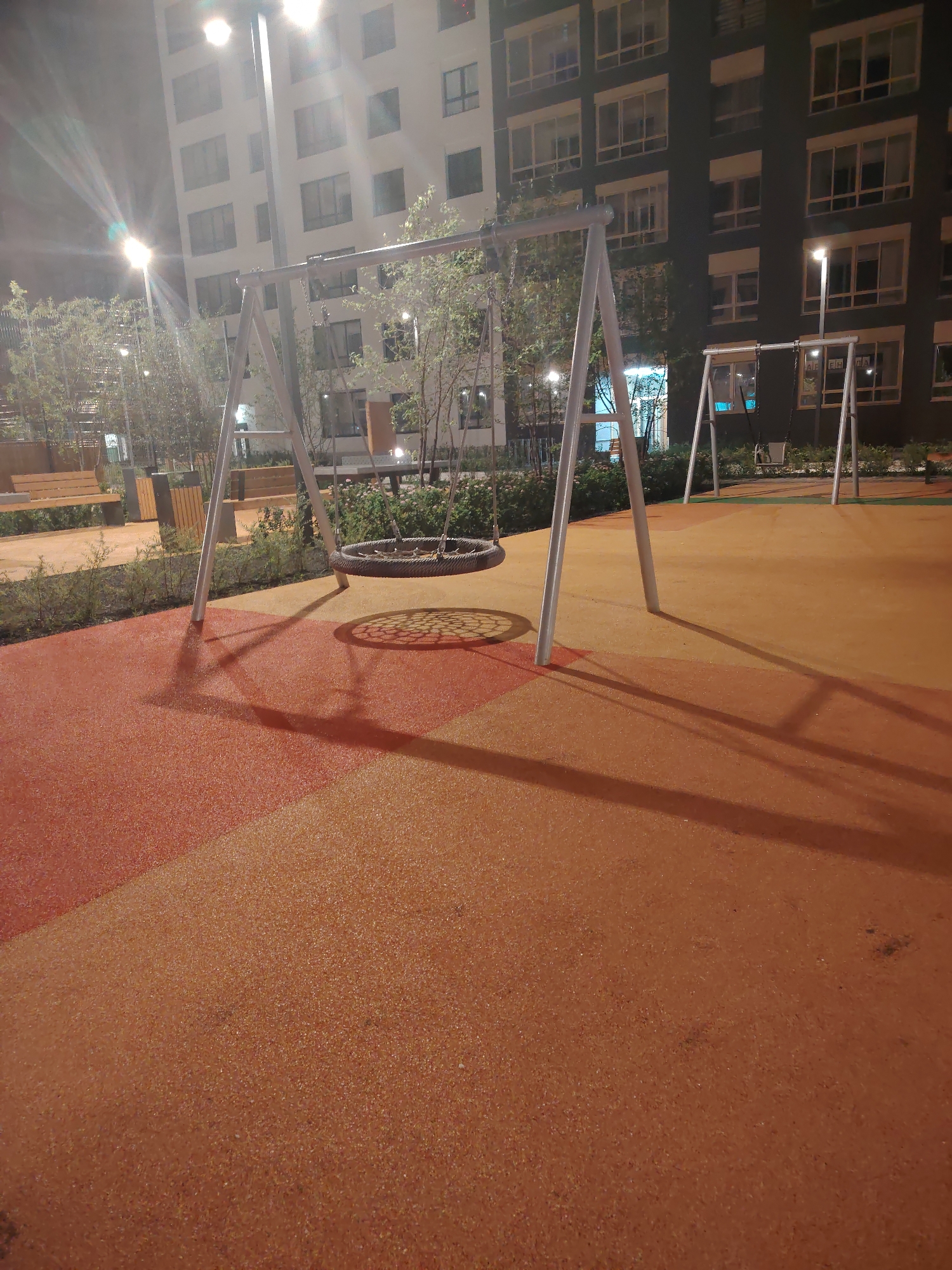 This is a low-angle, night-time photograph of a playground area. The foreground is dominated by a textured, rubberized surface, colored in two distinct sections: a reddish-orange on the left and a darker, more muted orange on the right. A prominent swing set with two tire swings is centrally located. The left swing, a circular tire, is closer to the camera, casting a distinct shadow on the ground. To the right, a second, smaller tire swing hangs from another swing set structure.
Behind the playground equipment, a line of low, green bushes runs horizontally across the image. Beyond the bushes, several tall, modern apartment buildings are visible. These buildings are multi-story structures with numerous windows. Streetlights illuminate the scene, creating bright spots and casting long shadows from the swing set and bushes onto the playground surface. The overall impression is of a well-maintained, urban outdoor recreation space at night. This scene is located in Yekaterinburg, Russia.