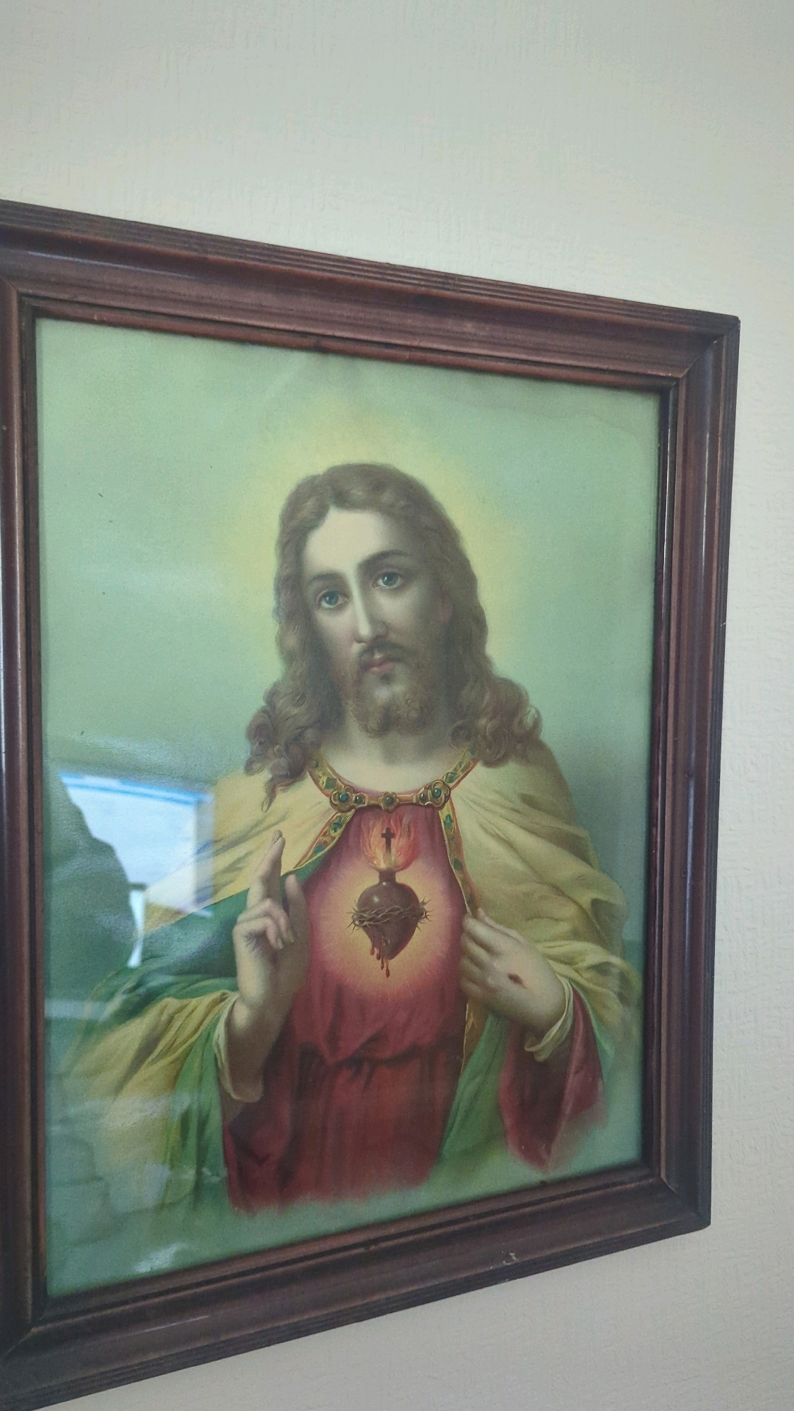 A framed painting of the Sacred Heart of Jesus hangs on a textured, light-colored wall. The painting, encased in a dark wooden frame, depicts Jesus with long, wavy brown hair and a beard. He has blue eyes and is looking forward. He is wearing a red robe and a yellowish-green garment draped over his shoulders. His right hand is raised, with two fingers extended in a gesture of blessing. His left hand is on his chest, over a prominent, glowing heart adorned with a crown of thorns and a flame. There are some reflections on the glass surface of the painting, including a blue and white rectangular shape on the left. The scene is dimly lit. The location of this painting within the city of Cornelscourt, Ireland, is not specified.