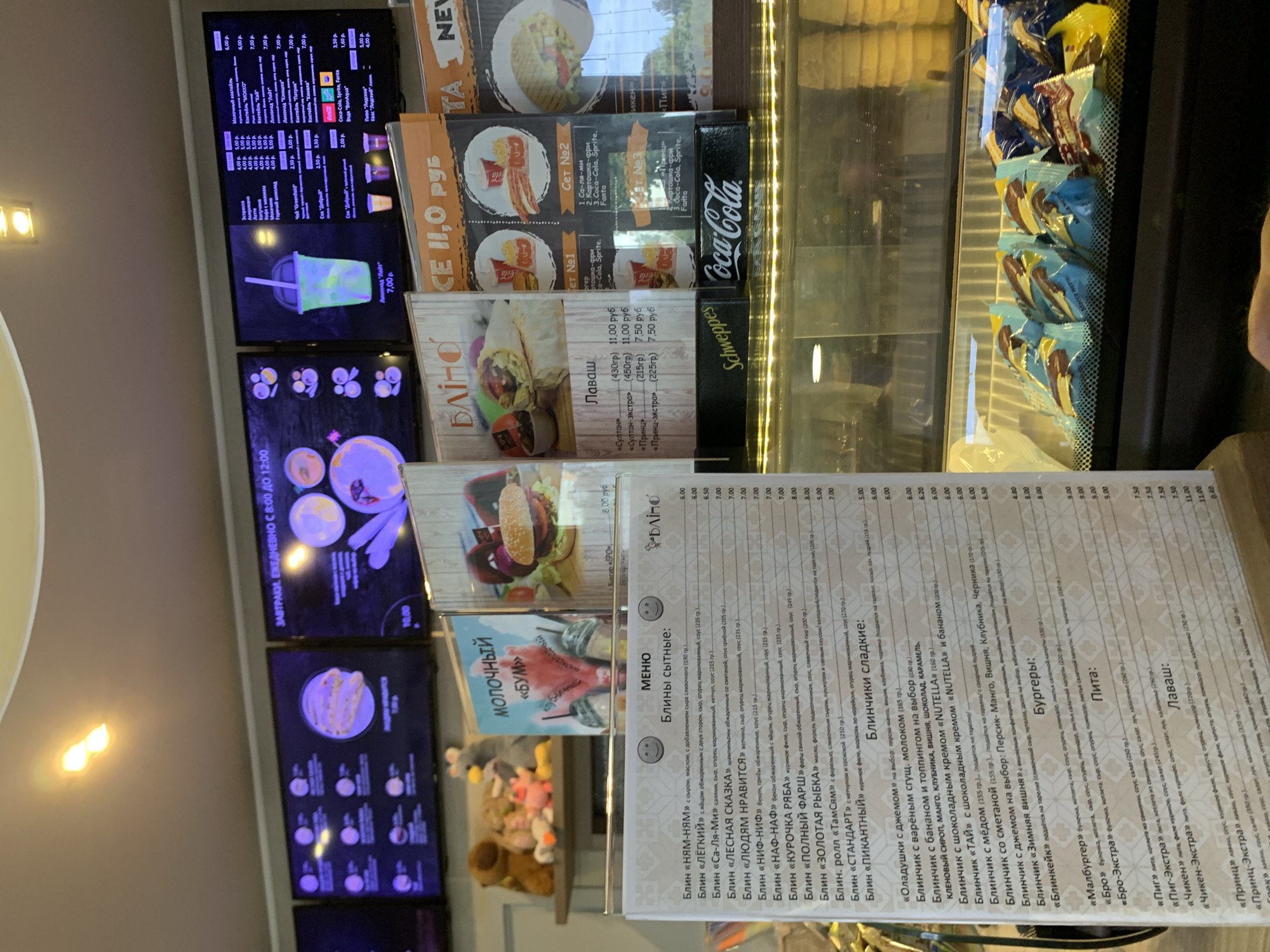 The image shows the interior of a fast-food establishment, likely a cafe or diner, in Sluck, Belarus. The establishment features several digital menu boards displaying various food and drink options. In the foreground, a printed menu is visible, listing items such as blini (pancakes), burgers, pita bread, and lavash wraps. The menu items are priced in Russian rubles. There are also display cases containing packaged food items, possibly desserts or snacks. The overall atmosphere appears casual and is designed to cater to customers seeking quick meals. The menu boards display images of food and prices, and the digital screens suggest a modern approach to menu presentation. The decor is simple, with emphasis on displaying the food offerings clearly. The background shows a blurry view of shelves with plush toys, suggesting a playful or family-friendly environment. The establishment seems to be open for business, with various food items displayed and ready for purchase.