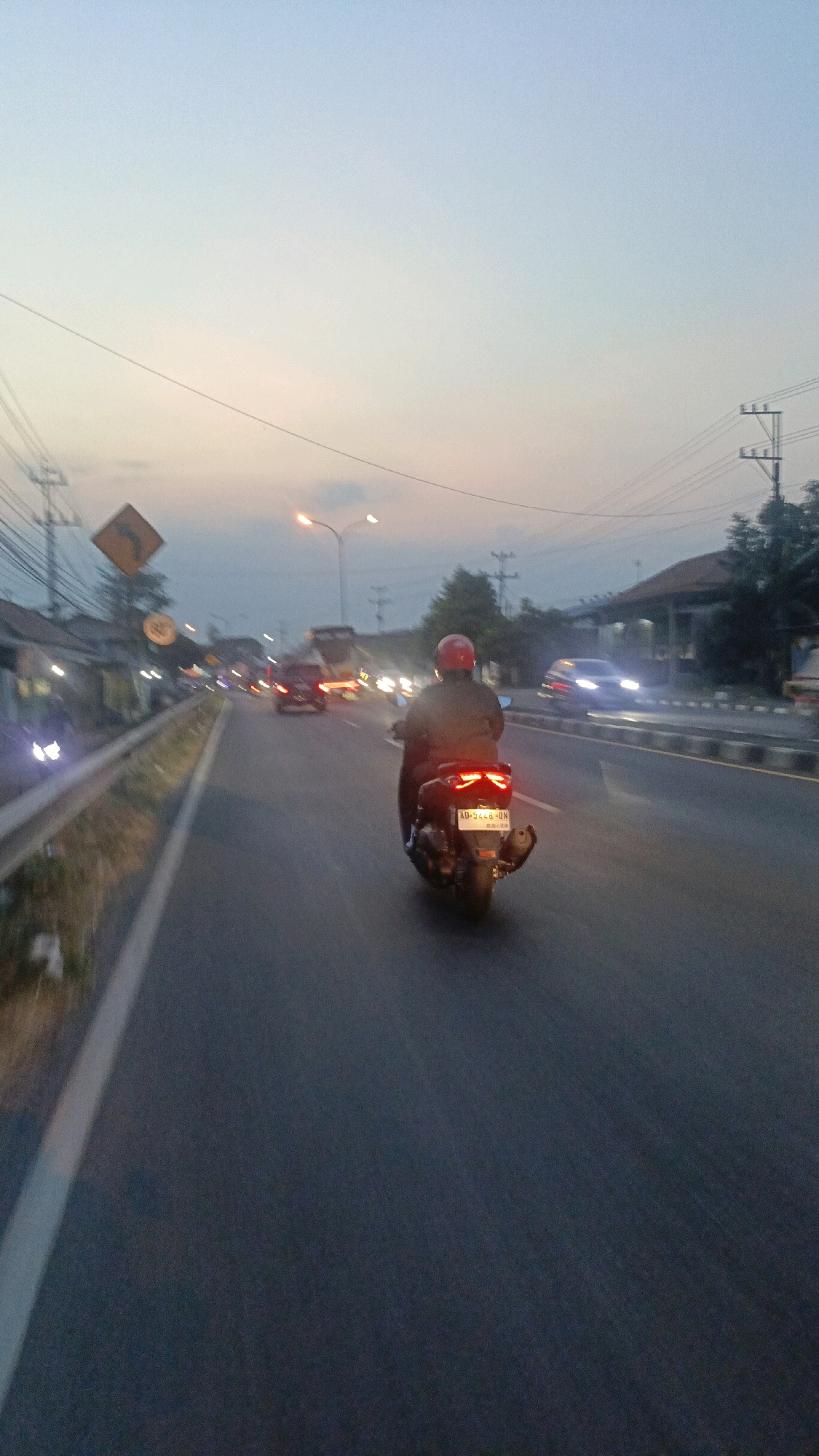 A perspective shot from the back of a motorcycle shows a road with several vehicles and streetlights in Banyudono, Indonesia. The sky is a pale blue, indicative of dusk or dawn. A person wearing a red helmet rides a motorcycle with its brake lights illuminated, facing away from the camera and driving on the right side of the road. Other vehicles, including cars and trucks, are visible in the distance, with their headlights and taillights creating streaks of light. The road is paved and appears to be a multi-lane highway with a barrier on the left side. Utility poles and power lines line the side of the road, along with some buildings and vegetation.