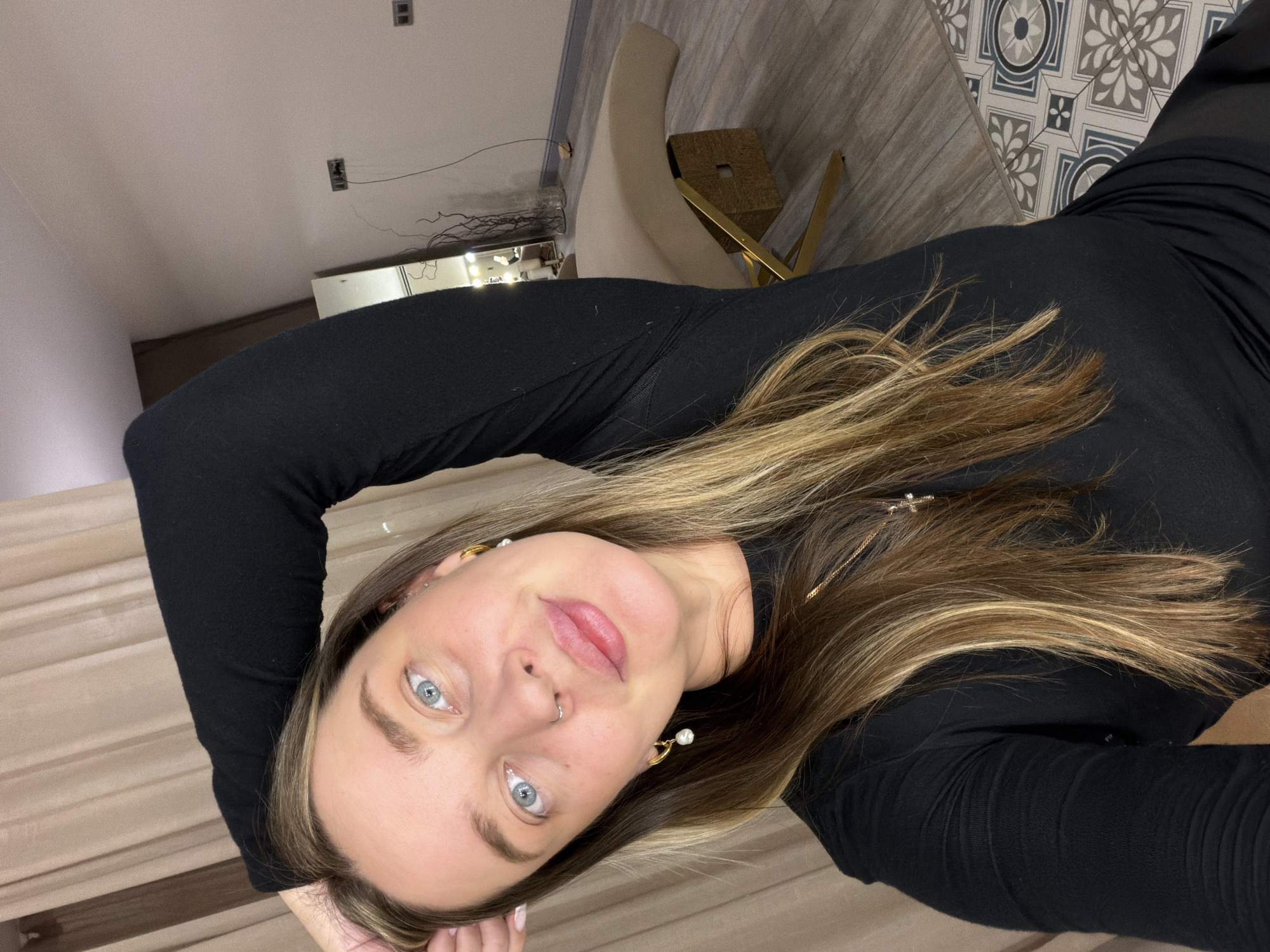 A portrait shot of a woman with long blonde and brown hair, wearing a black long-sleeved shirt. She has fair skin, blue eyes, and is wearing earrings and a nose ring. The background includes a beige chair, a patterned tile floor, a mirrored vanity, and light-colored curtains. The location is in the city of Unknown, Unknown.