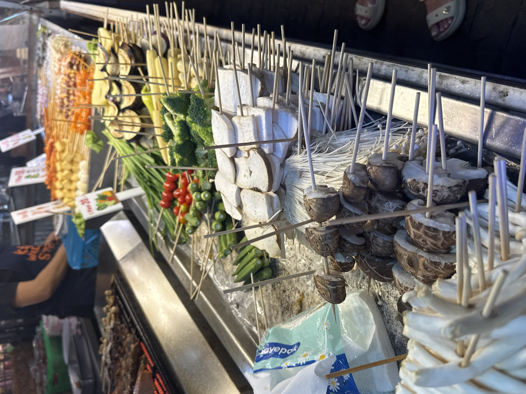 This media file captures a close-up view of a food stall, likely an outdoor market, in Karon, Thailand. The scene is dominated by an array of skewers filled with various food items, suggesting a street food vendor preparing for or actively serving customers.
The primary subjects are the food items themselves, presented in a visually appealing manner. We see skewers of colorful ingredients, including what appear to be processed meats or seafood in orange and red hues, interspersed with pieces of tofu. Further into the display, there are skewers of fresh vegetables such as broccoli florets, green beans, and cherry tomatoes. Notably, there are also skewers of sliced mushrooms and what looks like enoki mushrooms, as well as slices of eggplant and corn on the cob. The food is arranged neatly on wooden skewers, ready for grilling or other preparation methods.
Behind the food, partially visible, is a person, likely the vendor, wearing a dark t-shirt. Their arm is extended, and they are holding a blue bag, possibly for packaging purchased items. In the background, the feet of two individuals are visible, wearing sandals, suggesting the presence of customers or passersby. The overall setting appears to be a bustling market or street food area.
Notable details include the lighting, which suggests it could be daytime, possibly late afternoon or evening given the warm glow. The overall atmosphere seems lively, typical of a food market. There is some visible text on labels attached to the skewers, but it is not clearly legible. One label has an image of vegetables and text that is too blurry to read. Another label on a plastic bag reads "savepak".
The focus on the food and the implied activity of preparing and selling it point towards a commercial food establishment. The arrangement and variety of skewers indicate a popular street food offering, likely intended for takeaway.