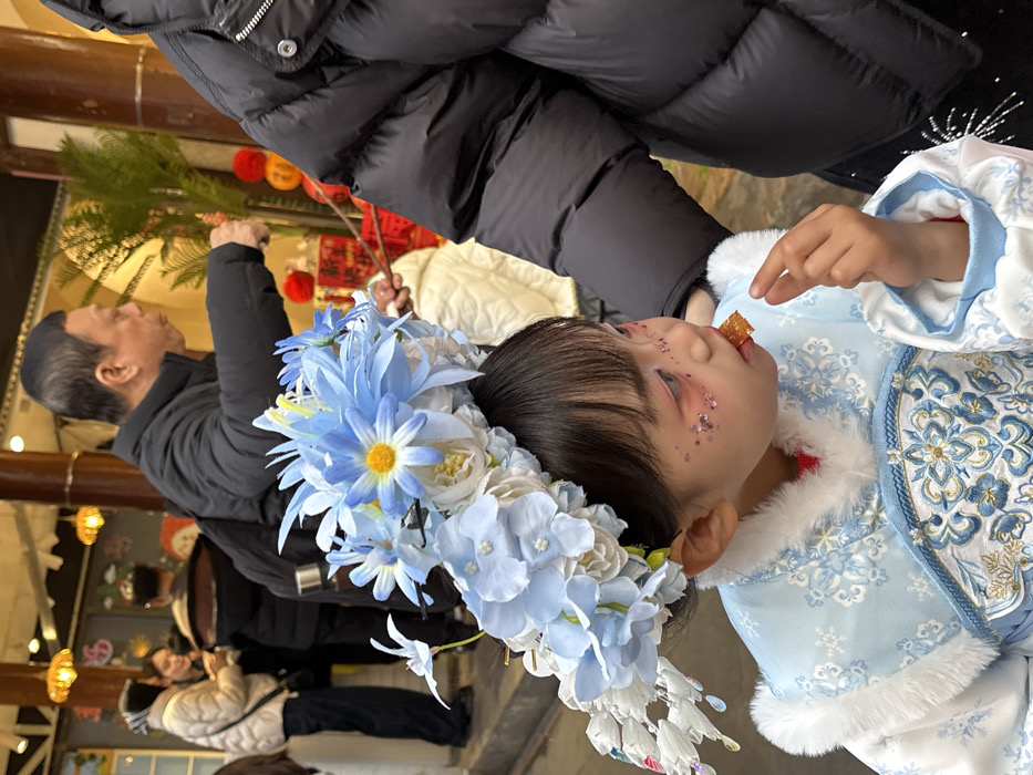 A young child, presumably a girl, is depicted in a medium-close-up view within a public indoor setting in Yuzhong District, China. She is positioned in the lower-left quadrant, wearing a light blue traditional-style garment featuring white fur trim and elaborate blue and white embroidery on the bodice. A large, complex headpiece, constructed from artificial blue and white flowers, including daisies and hydrangeas, adorns her head. Decorative elements, such as white conical shapes and reflective teardrop beads, extend from the left side of this headpiece. Her face is in profile, gazing slightly downward and to the right. She has visible makeup, including pink shading on her eyelids and glitter applied to her cheeks. She is holding a piece of golden-brown food, possibly a candied snack, in her right hand and appears to be consuming it. To her immediate right, the left arm and torso of an individual wearing a black puffer jacket are visible. In the mid-ground, a man with short dark hair, also in a black puffer jacket, is seen in profile, looking to his right while holding a thin stick with an object attached. The background consists of blurred figures, architectural wooden columns, a green potted plant, red lanterns, and two amber-colored pendant lights suspended from a ceiling, indicating a structured environment likely associated with a market or cultural venue.