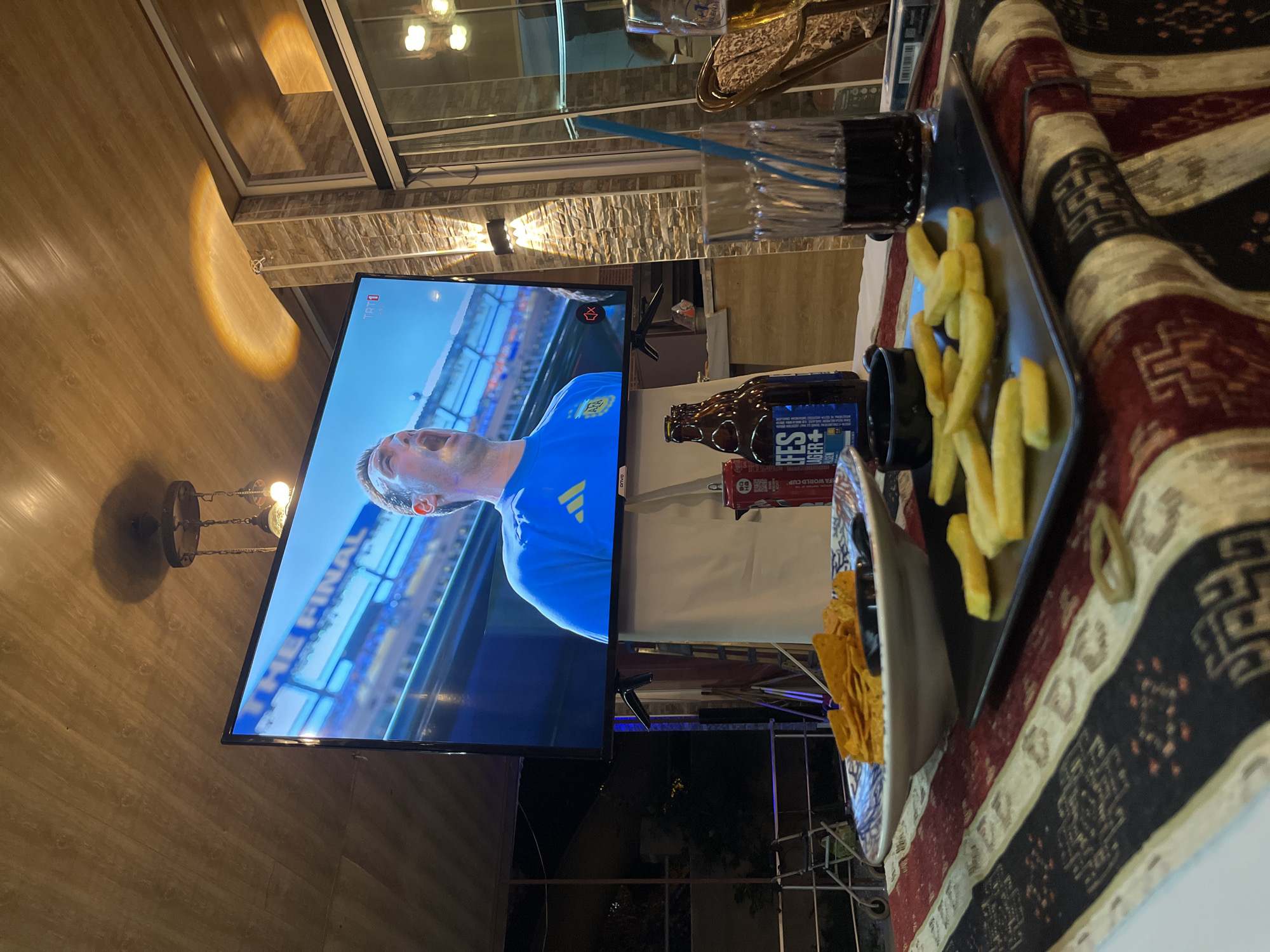 An outdoor seating area at a restaurant in Alanya, Turkey, is depicted, featuring a large television screen displaying a soccer match. On the table, there are snacks including tortilla chips and french fries, along with drinks and a bottle of Efes beer. The atmosphere suggests a casual viewing experience, possibly during a sporting event.