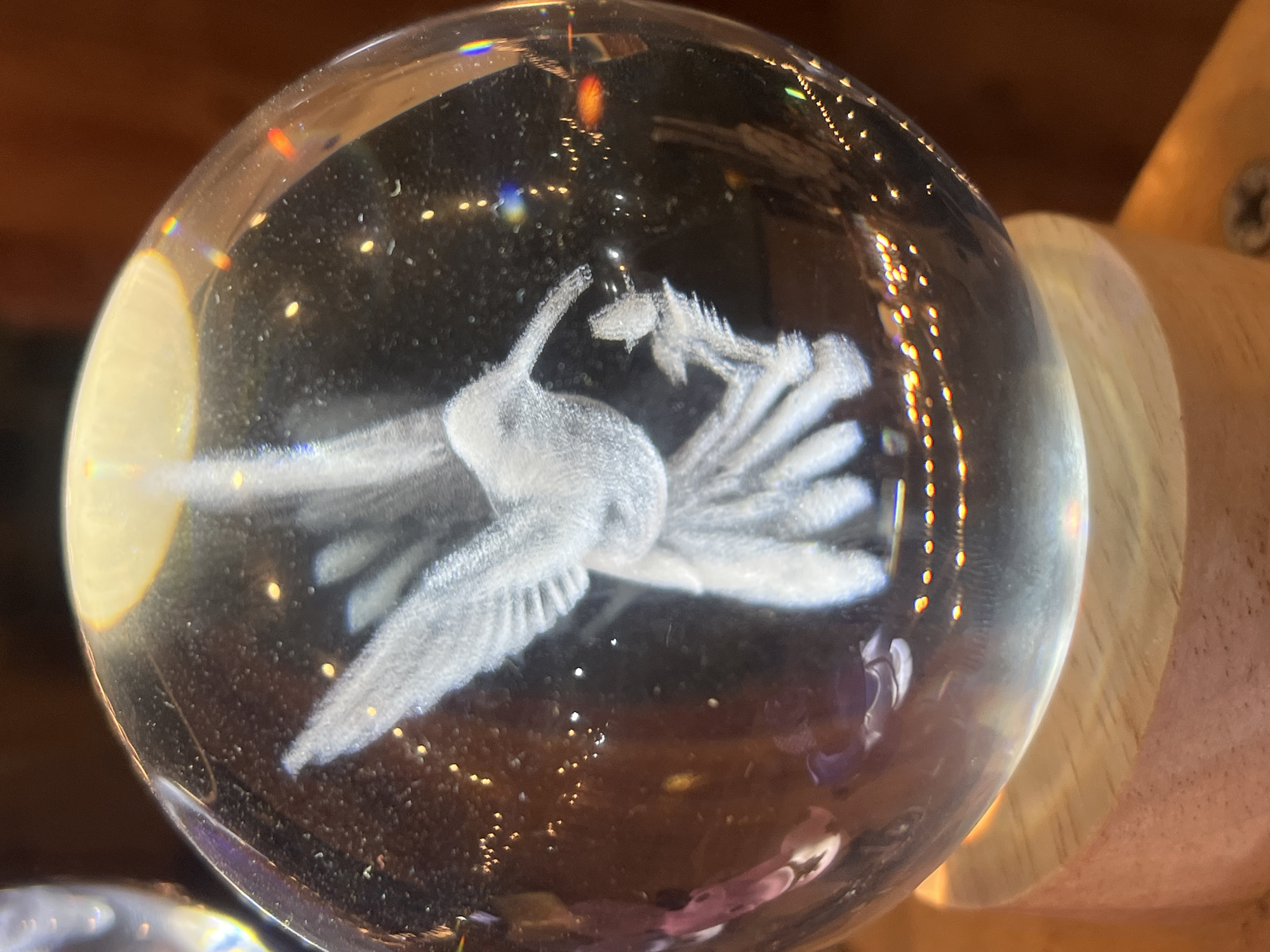 A clear, spherical glass paperweight with an image of a crane inside is positioned near a light-colored wooden object, potentially a support or base. The paperweight displays internal refraction and reflections, creating a glittering effect within the glass. The image of the crane is opaque white, contrasting with the transparent glass. The background appears to be a dark brown wooden surface. The specific location is unknown.