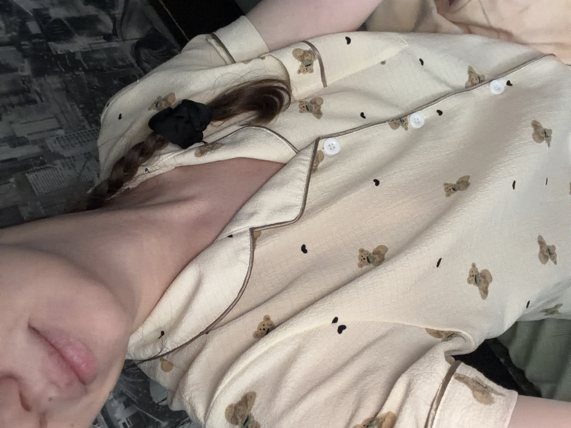 A close-up, medium shot shows a person's upper body, wearing light yellow pajamas with a teddy bear print. The pajamas have a collar and are buttoned up, with white buttons visible. A black scrunchie is holding back a braid of the person's brown hair. The background appears to be a wall with a city skyline pattern. The lighting is dim, and the image is slightly blurred. The location is not identifiable from the image.