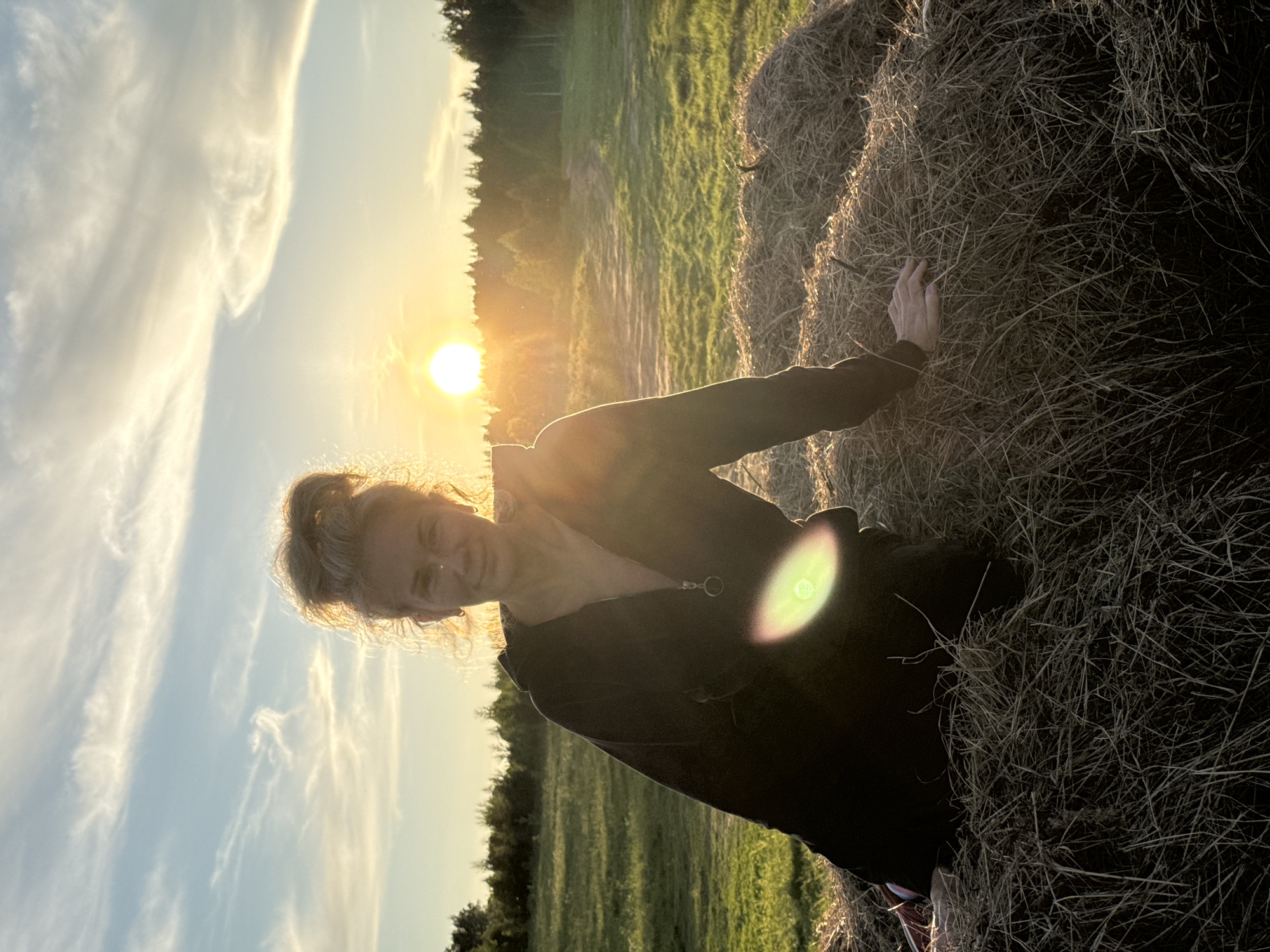 An outdoor portrait captures a woman during sunset in a rural area, likely near Стеклянный, Russia. The woman, with fair hair styled in an updo and a gentle smile, is positioned in the foreground, illuminated by the warm, golden light of the setting sun. She is wearing a dark, possibly zipped, top and her left hand is casually resting on a large, round hay bale to her right.
The sun, a brilliant, circular orb, is low in the sky directly behind her, creating a strong backlighting effect that silhouettes her figure and highlights the edges of her hair. A prominent lens flare, appearing as a bright, circular glow with rainbow-like rings, is visible over her right side.
The background features a verdant field or pasture stretching towards a distant line of trees or a forest along the horizon. The sky is bright with soft, wispy white clouds scattered across a light blue expanse, indicative of clear weather. The combination of the golden hour light, the natural setting, and the woman's relaxed expression evokes a sense of tranquility and contentment. There is no visible text in the image.