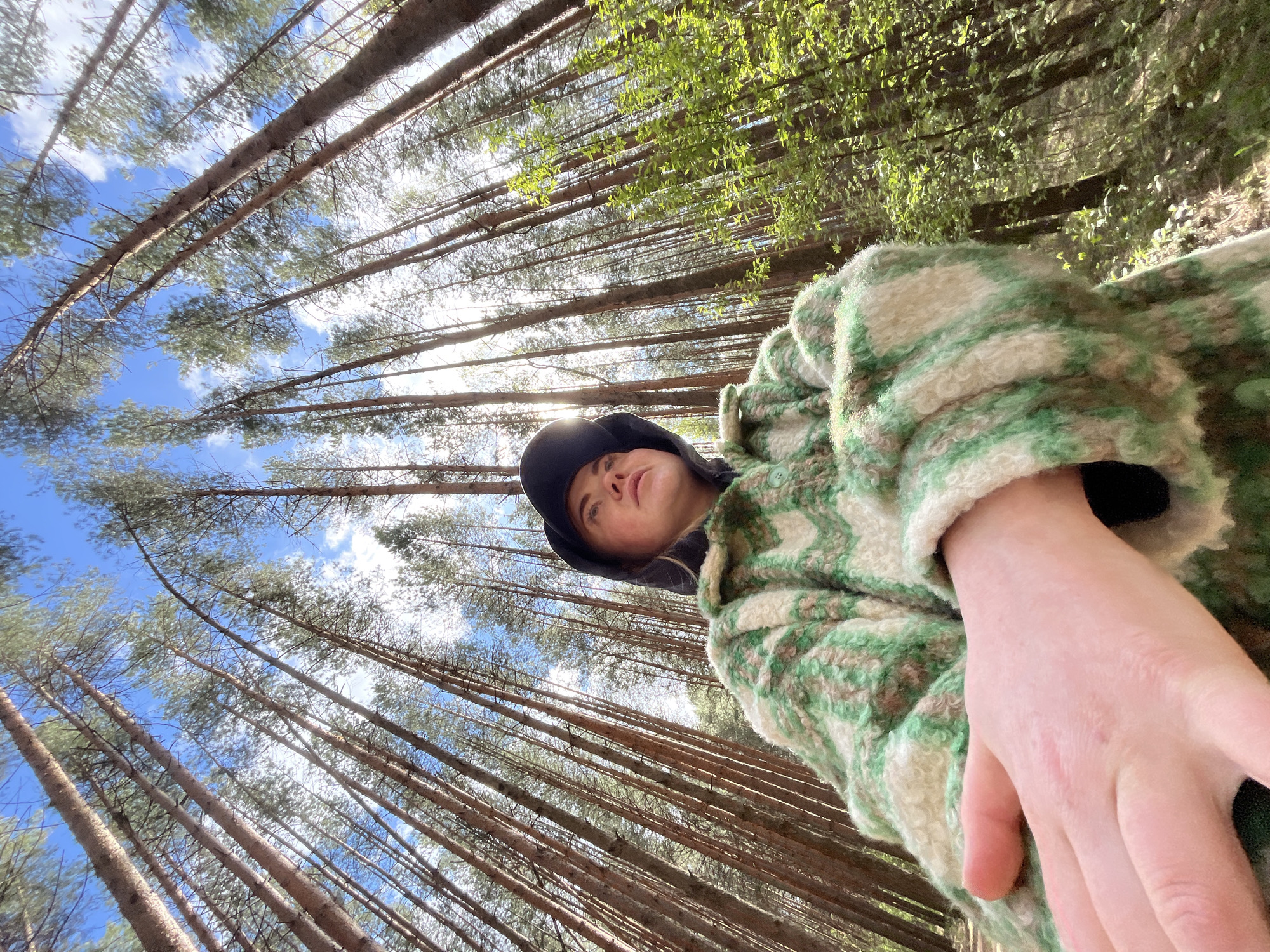 The image depicts a person wearing a green and beige jacket and a black cap, looking up at the camera. The person is situated in a forest with tall, straight trees stretching upwards, creating a converging perspective. Glimpses of blue sky and white clouds are visible through the tree canopy. The location is likely a forest, and given the city information, a forest near Chaikovsky, Russia.