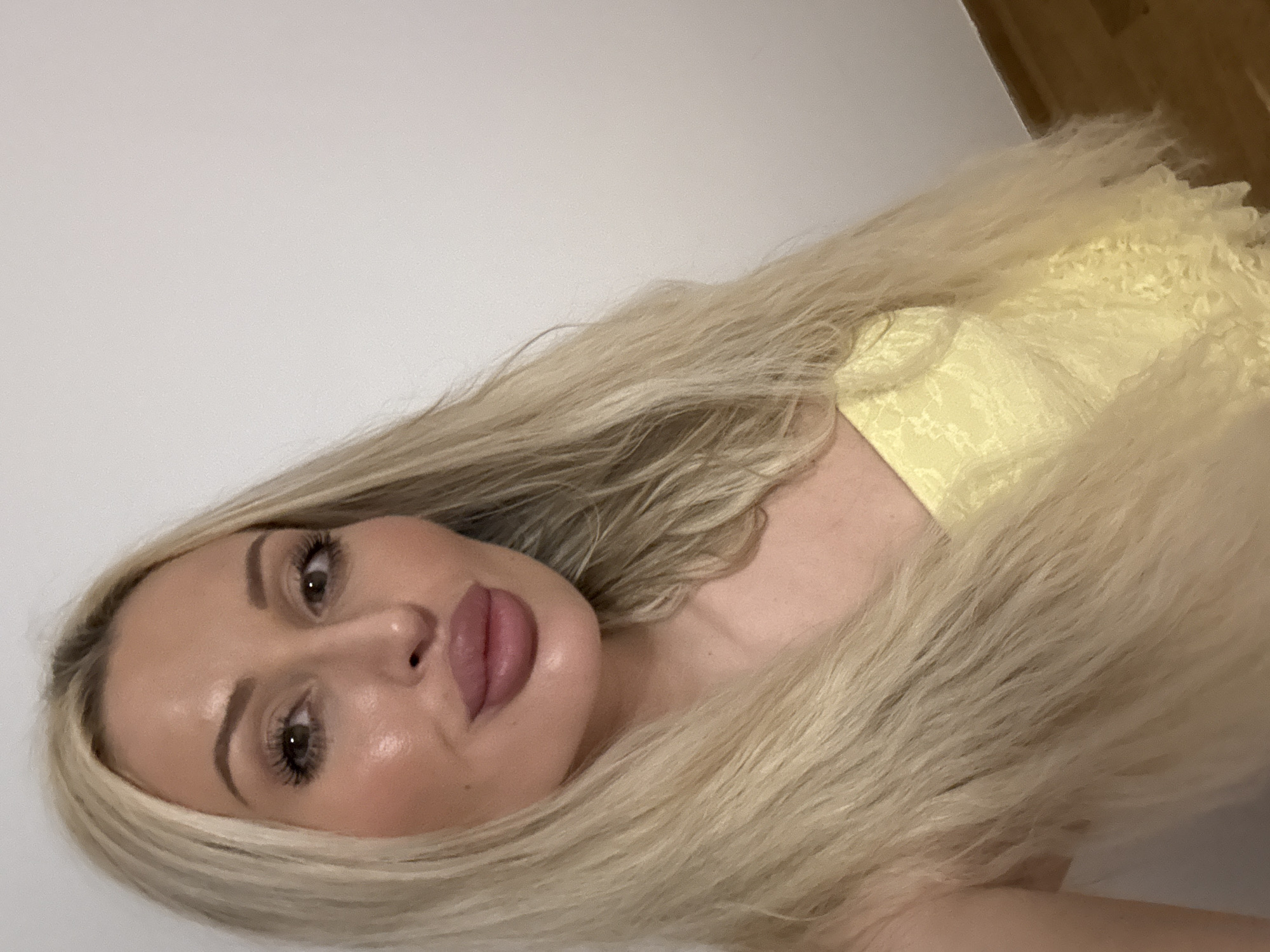 The media file is a close-up, waist-up, indoor portrait of a young Caucasian woman with long, blonde hair. She is wearing a yellow, lace-detailed garment. The woman is positioned with her head tilted, looking directly at the camera. Her eyes are open, and her lips are slightly parted, suggesting a neutral or possibly alluring expression. She has visible makeup, including eyeliner and lipstick. The background is a plain, light-colored wall, with a hint of wooden furniture visible in the upper right corner, indicating an indoor setting. The lighting appears to be artificial, creating a soft glow on her skin. No specific activity or event is evident; the focus is on the subject's portrait. There is no visible text in the image. The overall impression is that of a casual selfie or posed portrait.