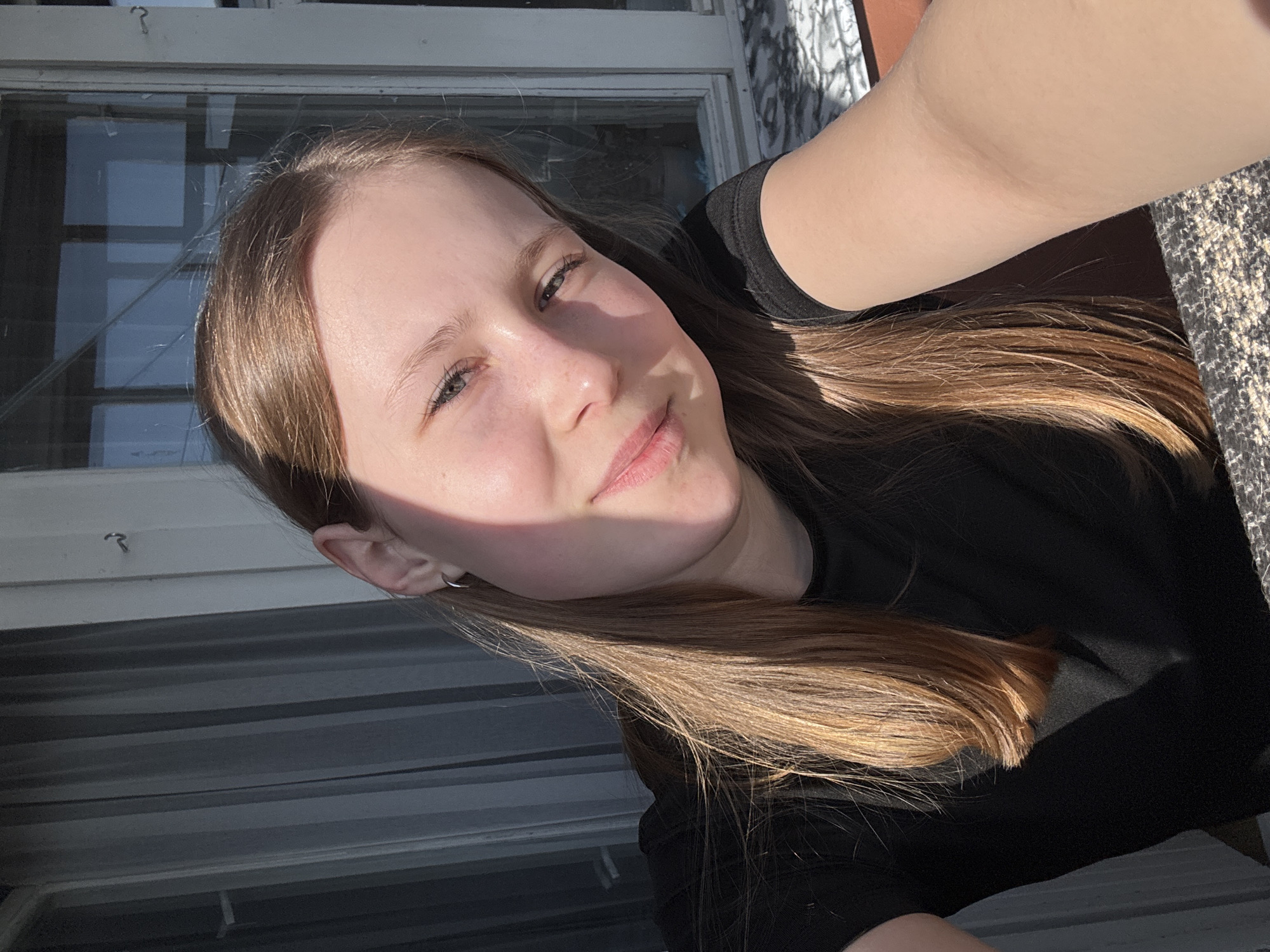 A young person with light brown hair is taking a selfie. The individual is wearing a black shirt and has a small hoop earring in their left ear. The background includes a window with multiple panes and an exterior wall. The location is Chaikovsky, Russia.