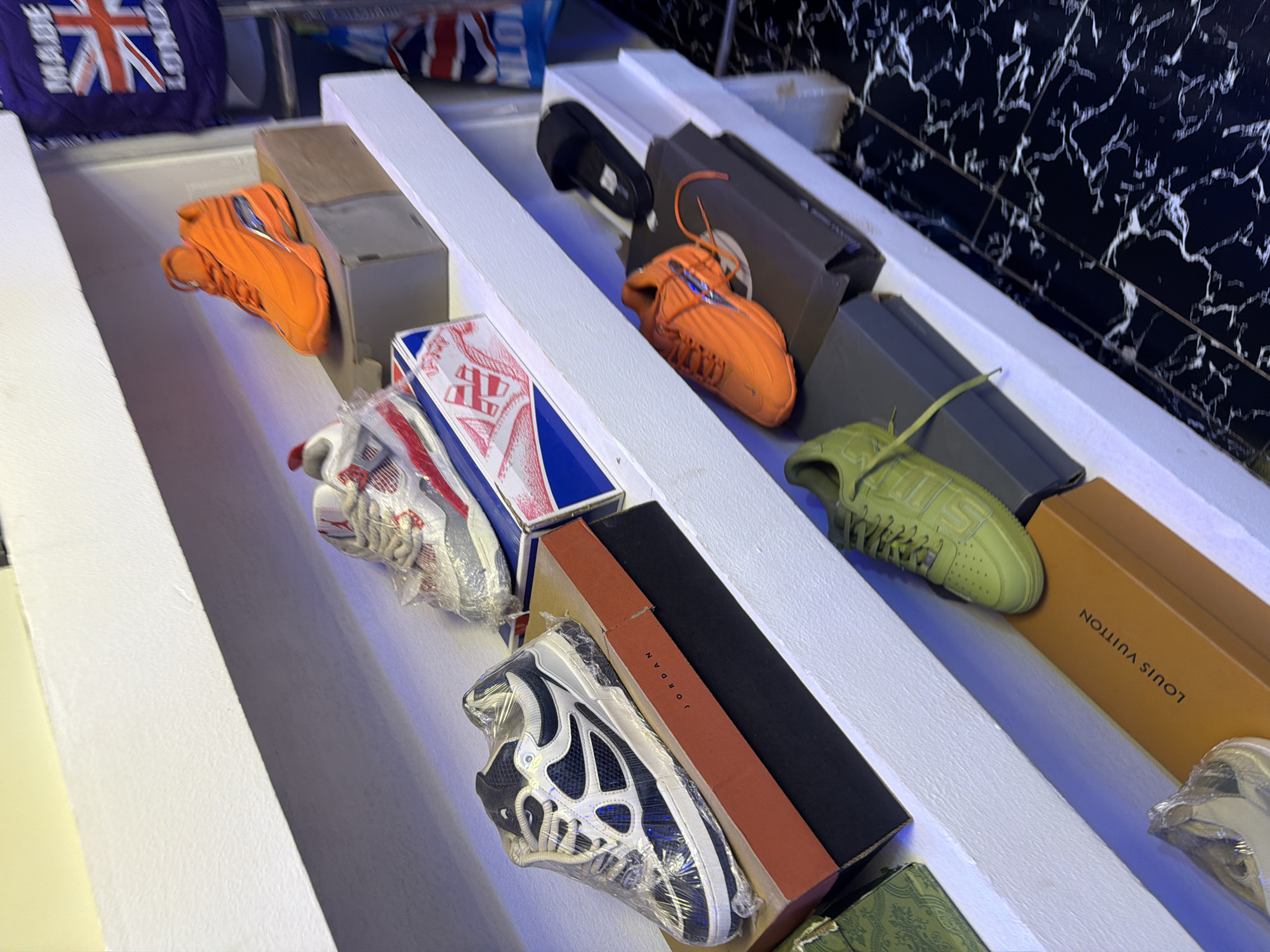 The media file displays a retail setting, likely a shoe store in Bukuru, Nigeria. The main subjects are various pairs of athletic footwear, neatly displayed on white shelves. Several pairs of sneakers are visible, including bright orange ones, a lime green pair with "SUPREME" branding, and a black and white pair wrapped in plastic with "JORDAN" visible on the box. Other objects in the scene include shoe boxes, one of which is a distinctive Louis Vuitton box. The background features a black and white marbled wall, adding to the modern aesthetic of the store. The lighting appears to be artificial, suggesting the image was taken indoors, possibly during the day or evening. There is no activity or event taking place, as the scene is static, showcasing merchandise. The overall impression is one of organized retail display.
