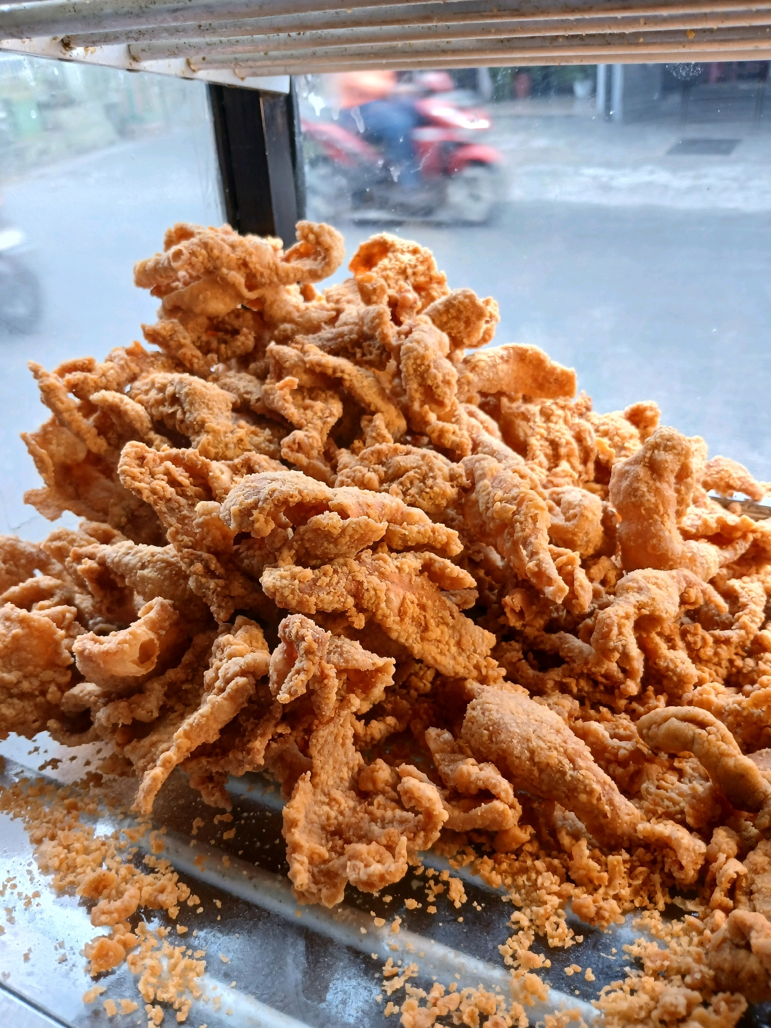 A close-up, overhead shot captures a heap of golden-brown fried chicken pieces piled high on a metal tray. The chicken pieces are irregular in shape, some appearing to be strips, while others are more curled and tubular. The coating on the chicken is textured and uneven, with some crispy bits visible. Small crumbs of the fried coating are scattered around the base of the pile and on the tray.
In the background, a blurred outdoor scene is visible through a glass or transparent surface, suggesting the food is displayed in a window or counter. Through the blur, a red motorcycle with a rider wearing a blue shirt is discernible, moving along a street. The overall environment appears to be an outdoor urban setting. The image does not provide explicit details to pinpoint the location within Depok, Indonesia, beyond suggesting a street-side food vendor or establishment.
