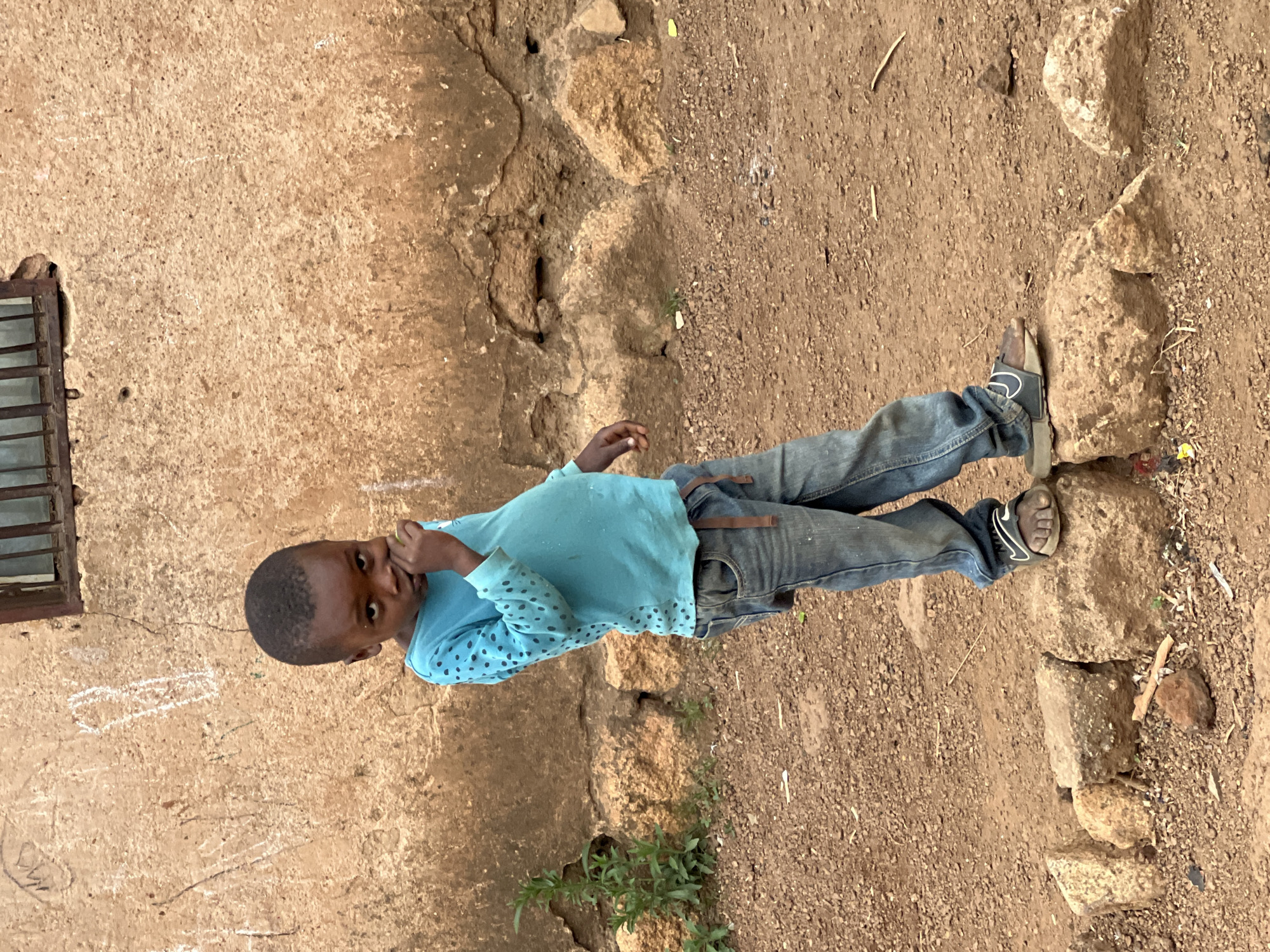 A young Black child stands near a wall, his right hand touching his face. He is wearing a light blue, long-sleeved shirt with dark polka dots and faded blue jeans with a brown belt. His feet are clad in dark sandals with a white Nike swoosh logo. He stands on a dirt and gravel ground interspersed with rocks, some forming a rough border. The weathered wall behind him is light brown and has a barred window. There appear to be chalk markings on the wall. The photo was taken in Jos, Nigeria.
