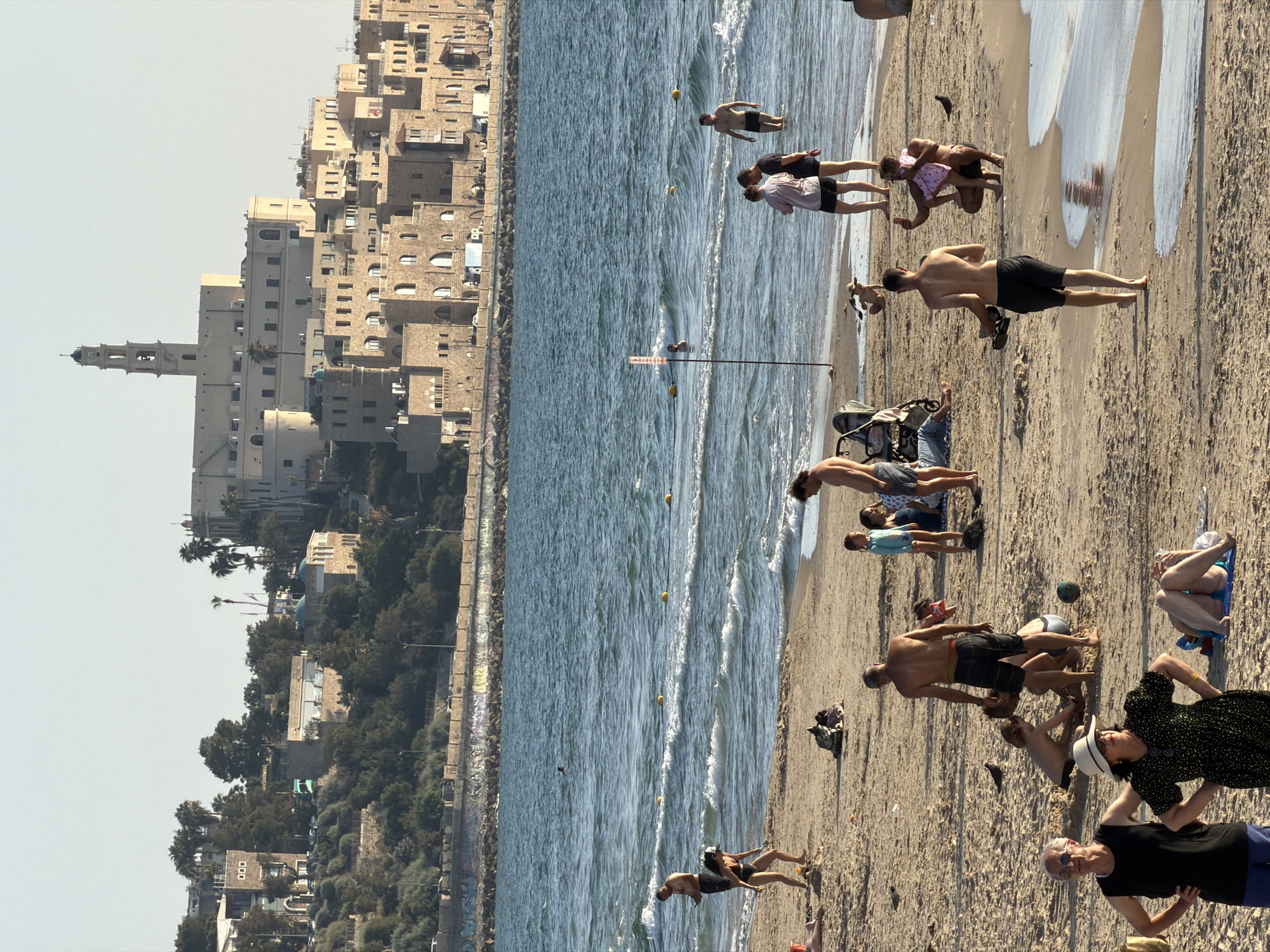 The image depicts a bustling beach scene on a sunny day, likely in Jaffa, an ancient port city that is now part of Tel Aviv-Yafo, Israel. In the background, the distinctive historical architecture of Jaffa, characterized by its stone buildings and a prominent clock tower, is visible against a clear sky. The foreground is dominated by a sandy beach populated by numerous people engaged in various activities. Some are sunbathing on towels, while others are wading or swimming in the shallow ocean waves. Children are visible playing in the sand, and some adults are walking along the shore. The sea itself is blue with white-capped waves, and yellow buoys mark a boundary in the water. The overall atmosphere is one of leisure and recreation, with a mix of individuals and groups enjoying the beach and the sea.