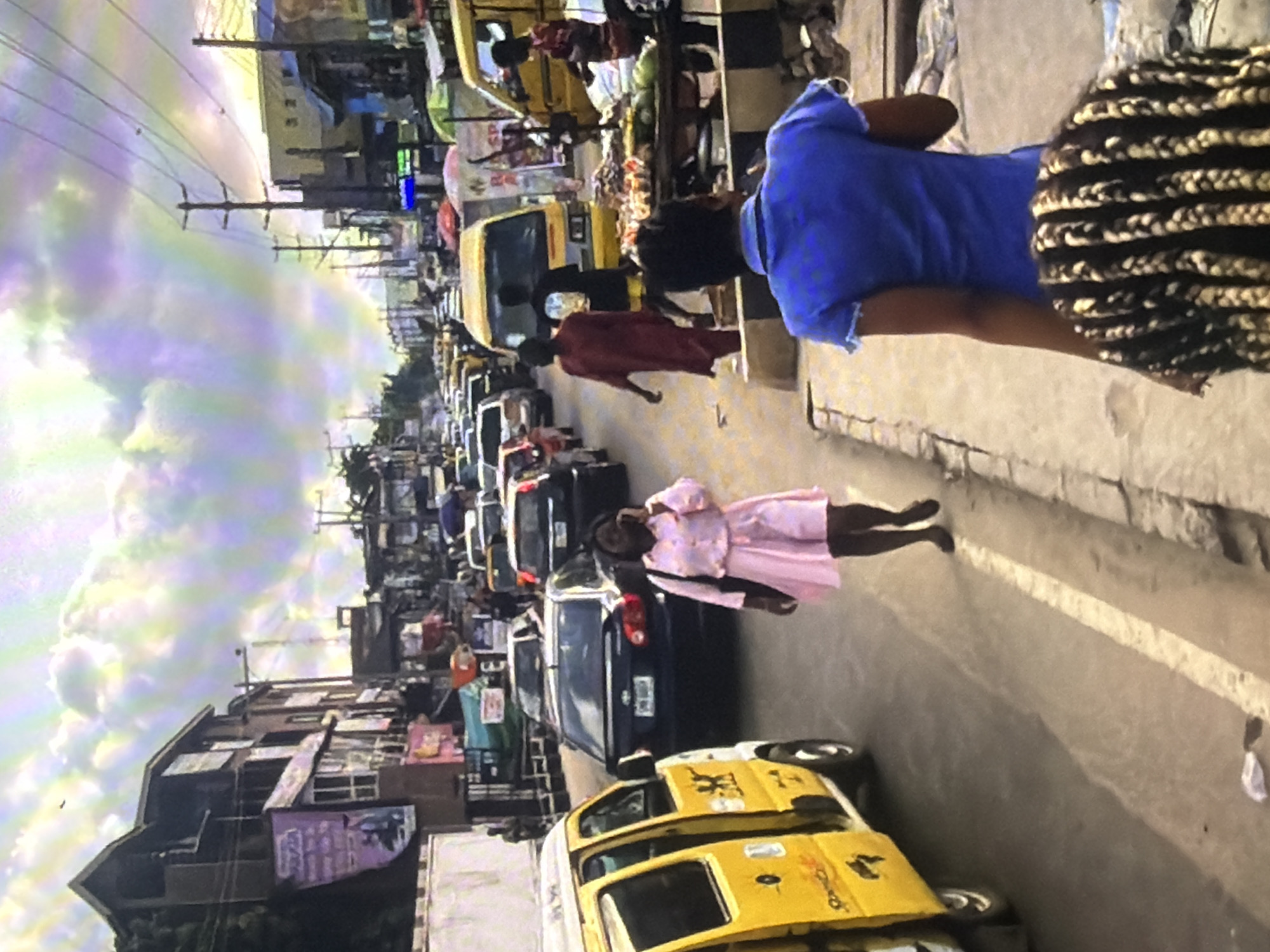 The image depicts a busy street scene in Bukuru, Nigeria, with a notable traffic jam. Yellow minibuses, a common mode of transport in Nigeria, are visible, along with other cars. Pedestrians walk along the side of the road, some engaged in conversations, while others navigate through the congestion. Buildings line the street, some displaying advertisements. The sky is filled with dramatic, colorful clouds, possibly indicating either sunrise or sunset, or unusual atmospheric conditions. A market stall with fruits and vegetables is set up on the roadside. The overall atmosphere suggests a vibrant, bustling urban environment.