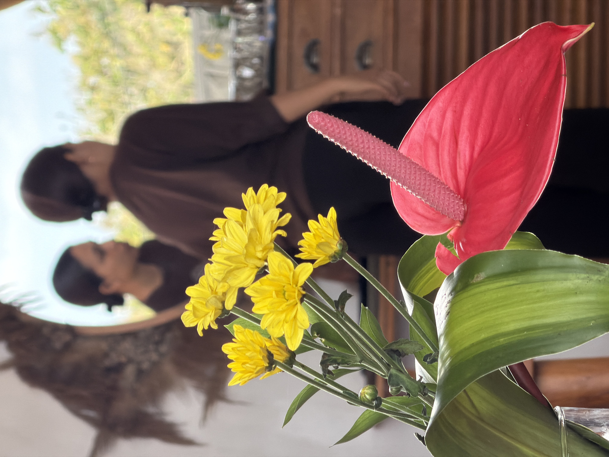 In Kuta Selatan, Indonesia, an indoor scene features a close-up view of a vibrant red anthurium flower and a cluster of yellow chrysanthemums in the foreground, placed in a glass vase. Behind the flowers, a woman with dark hair tied back is visible in the background, facing away from the camera and appearing to be in a room with wooden cabinets and a window that lets in natural light. The reflection of the woman and a decorative element, possibly a sculpture or dried foliage, is visible in a mirror on the wall to the left. The overall atmosphere is casual and natural.