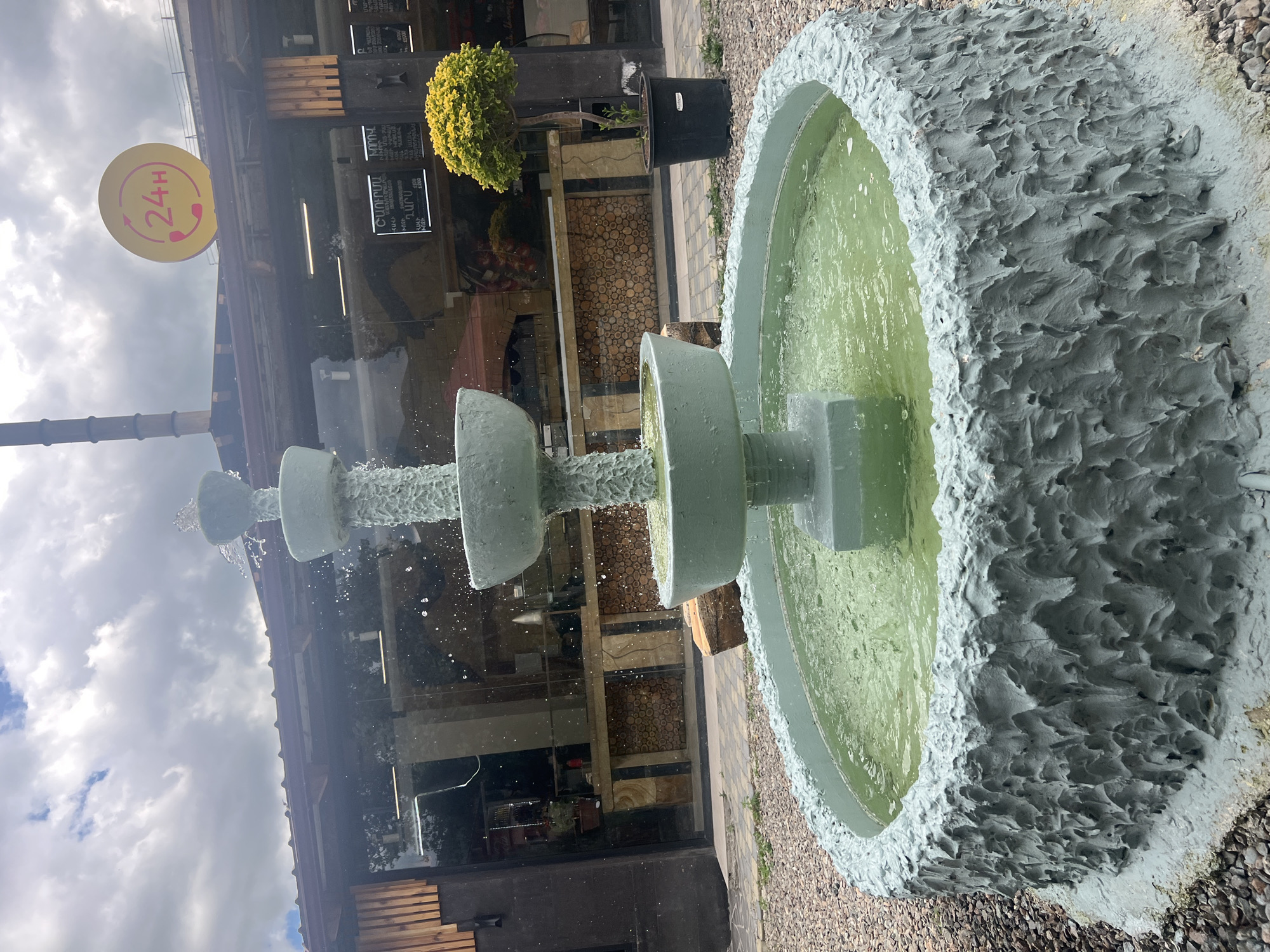 The media file shows a close-up, slightly angled shot of a tiered fountain. The fountain is made of a light gray, textured material and has water flowing down its levels. The water in the basin is a murky green color. The background is slightly out of focus but reveals a building with large windows and some greenery. To the left of the fountain, a yellow circular sign with a red outline and a telephone icon is visible. Inside the telephone icon, the text "24H" is written in red. The sign appears to be attached to a pole. Beyond the building, there is a cloudy sky. The overall impression is of an outdoor setting, possibly a cafe or restaurant, given the "24H" sign and the building's facade. The lighting suggests it is daytime, but the cloudy sky indicates overcast weather.