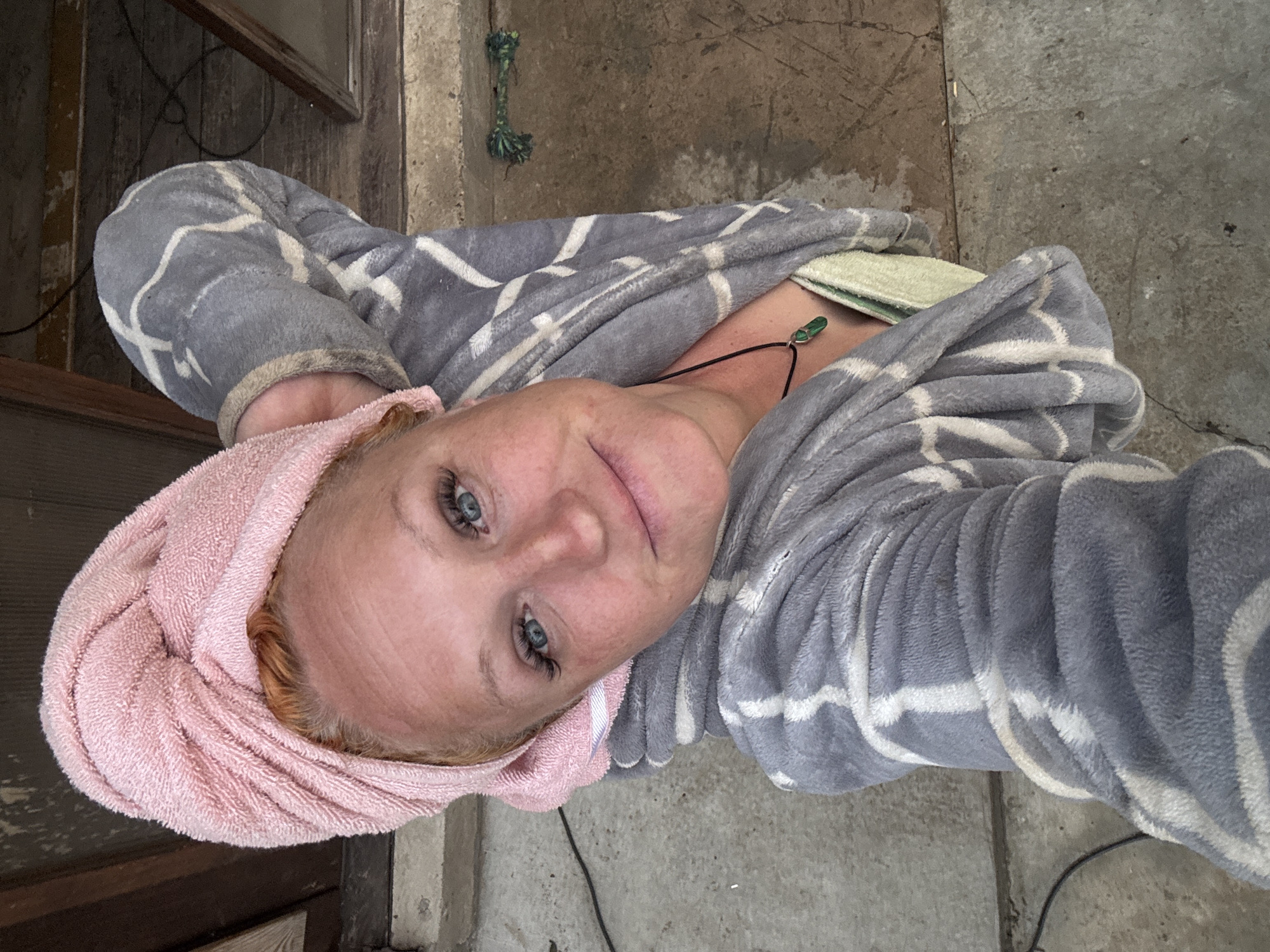 A selfie-style portrait shows a fair-skinned woman with blue eyes and reddish hair, wrapped in a pink terry cloth towel on her head. She is wearing a soft, grey and white patterned robe or blanket. A dark cord necklace with a small, green, elongated pendant rests on her neck. She looks directly at the camera with a calm, neutral expression.
The background suggests an outdoor or semi-outdoor setting. To the left, there are weathered wooden elements or a distressed wall, possibly part of a building or structure. The ground beneath her appears to be rough, grey concrete or paving, with visible cracks and inconsistencies. A thin, dark cable or wire runs across the concrete floor. In the upper background, a small, green, braided rope object hangs from the wall. The lighting indicates it is daytime. The overall scene conveys a casual and unpretentious atmosphere in Chaikovsky, Russia. No visible text is present.