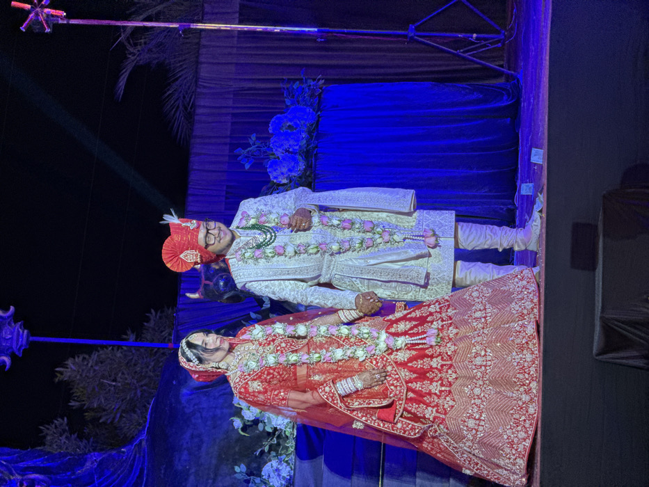 A bride and groom stand together at their wedding. The bride wears a red and gold lehenga, while the groom is in an off-white sherwani and red turban. Both wear floral garlands. They stand on a blue-draped stage, with blue flowers and greenery. A lighting rig is visible in the background. The overall image is vibrant with rich colors.