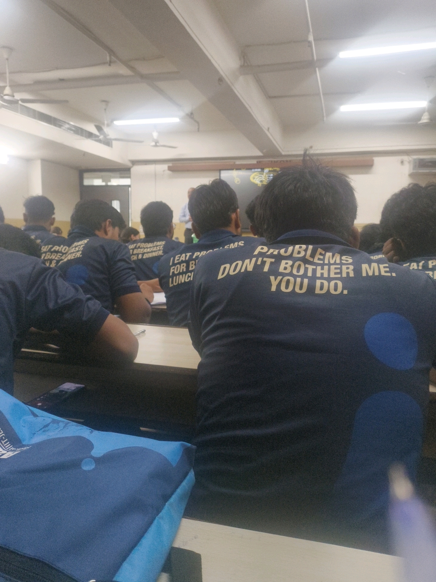 The image depicts an indoor scene, likely a classroom or lecture hall, in Kota, India. The perspective is from behind a group of students wearing dark blue t-shirts with yellow lettering. The t-shirts of several students have text that reads, "I EAT PROBLEMS FOR BREAKFAST, LUNCH & DINNER." One t-shirt in the foreground has the text, "PROBLEMS DON'T BOTHER ME. YOU DO." A person, presumably an instructor, is visible in the background, standing and facing the students. The room has a white ceiling with fluorescent lighting and ceiling fans. Desks and chairs are arranged in rows, with some students actively engaged in listening or taking notes. A blue bag with a graphic design is visible on a desk in the foreground. The overall atmosphere suggests an educational setting, with the prominent t-shirt slogans hinting at a specific student culture or event.