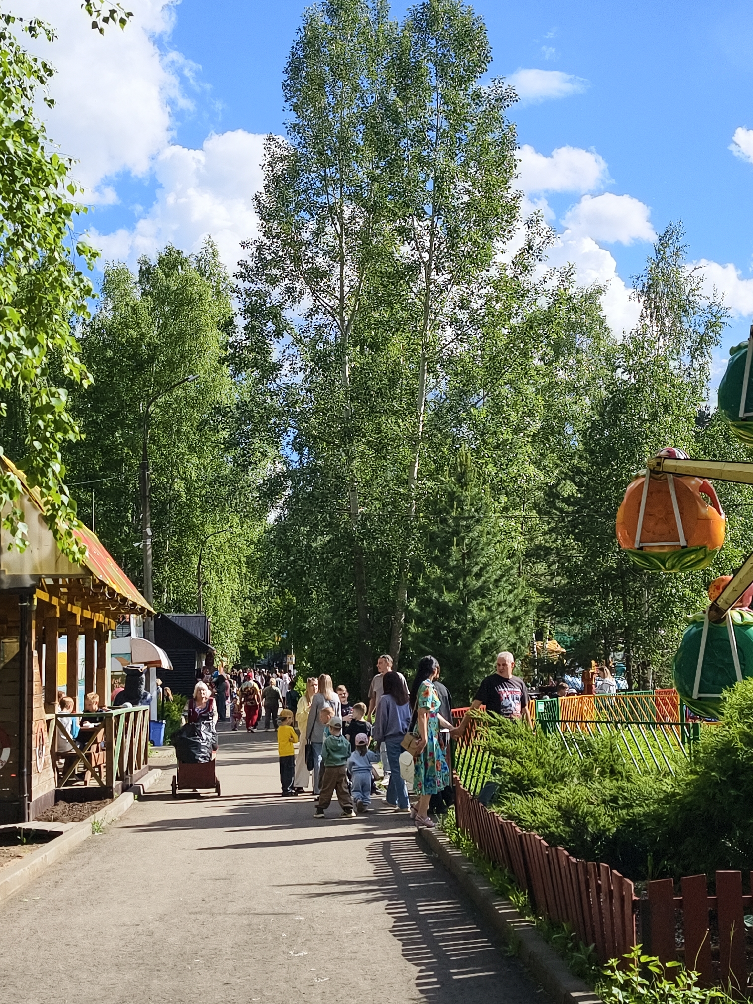 The image captures a sunny, crowded pathway within a park in Chaikovsky, Russia. The scene is filled with people of various ages walking along a paved path. A building sits to the left of the path and features an open-air dining area. To the right, is a fence separating the walking path from a carnival ride consisting of fruit-shaped carriages. The trees dominate the background. The sky is blue with fluffy white clouds.