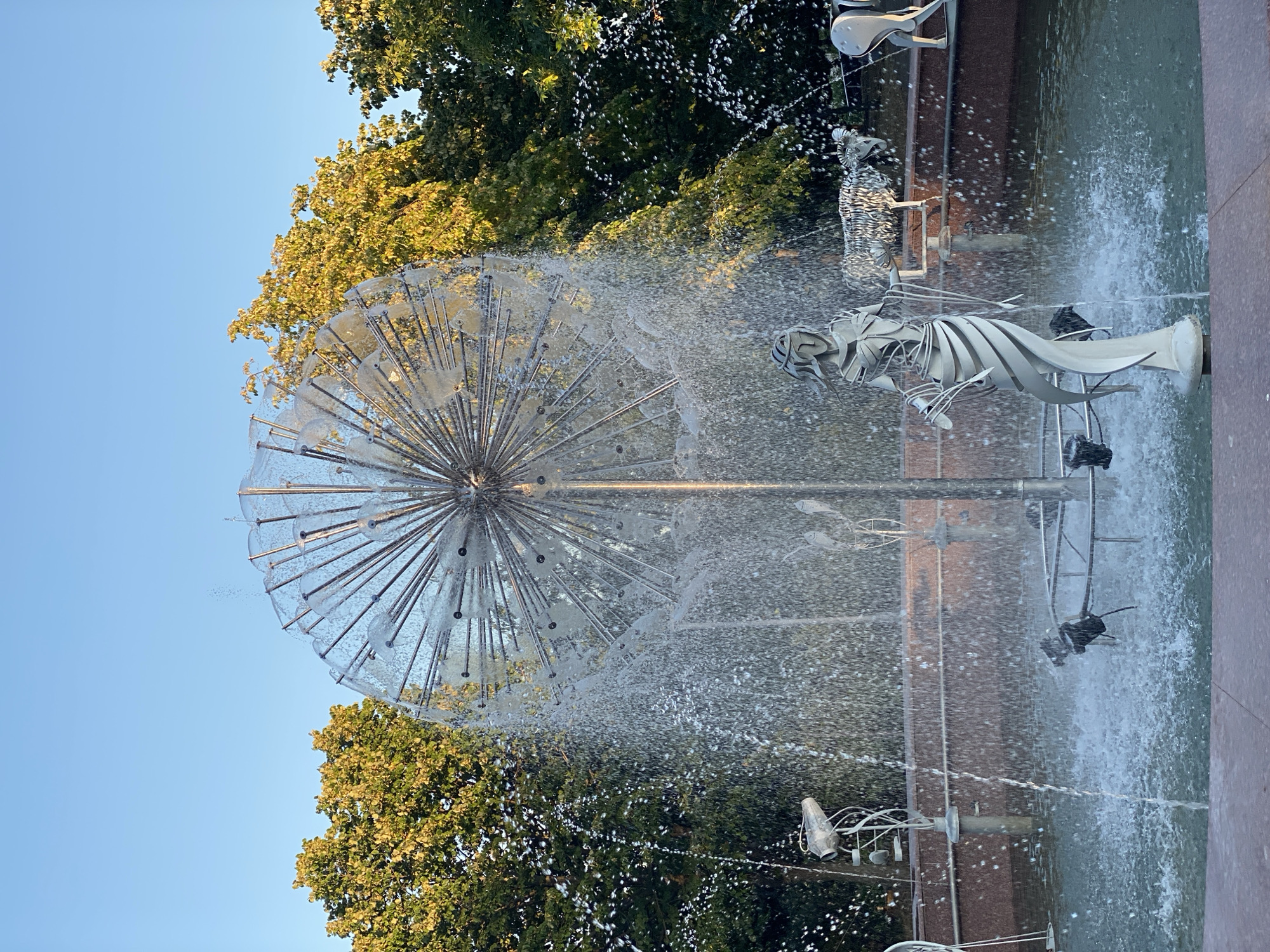 The image shows a fountain in a park, with a large, spherical water feature resembling a dandelion seed head. Water sprays out from the metallic structure, creating a shimmering effect. In front of the main water feature, there is a statue of a woman in a flowing gown, made of white metal. Behind the woman statue, to the right, are metal sculptures of animals, appearing to be sheep or goats. The fountain is surrounded by trees with green and yellow foliage, indicating it might be autumn. The sky above is clear blue. The fountain is situated in a park, likely in Chișinău, Moldova, as suggested by external context. The overall scene depicts a public space with artistic elements and nature.