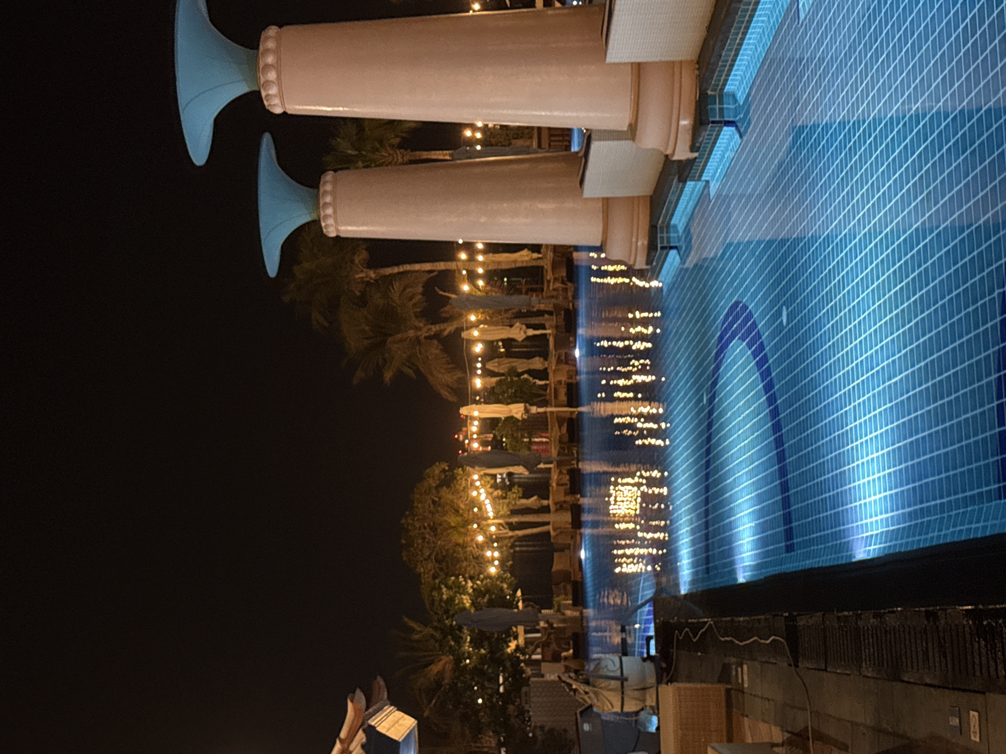 The image captures a nighttime scene at a poolside resort in Dubai, United Arab Emirates. Dominating the foreground are two large, light pink, fluted columns topped with bright turquoise cone-shaped accents, indicative of a grand architectural style. The columns sit at the edge of a swimming pool, which is illuminated from below, casting a vibrant blue glow onto the water's surface and its tiled floor. The pool reflects a string of warm yellow fairy lights that hang above a line of palm trees and lounge chairs in the background, creating a festive and inviting atmosphere. Several closed, light-colored umbrellas are visible near the lounge area, suggesting the resort is prepared for daytime use. A few shadowy figures can be faintly discerned near the edge of the pool on the left, suggesting the presence of patrons or staff, though they are not clearly defined. The overall ambiance is one of luxury and relaxation, characteristic of Dubai's upscale leisure establishments.