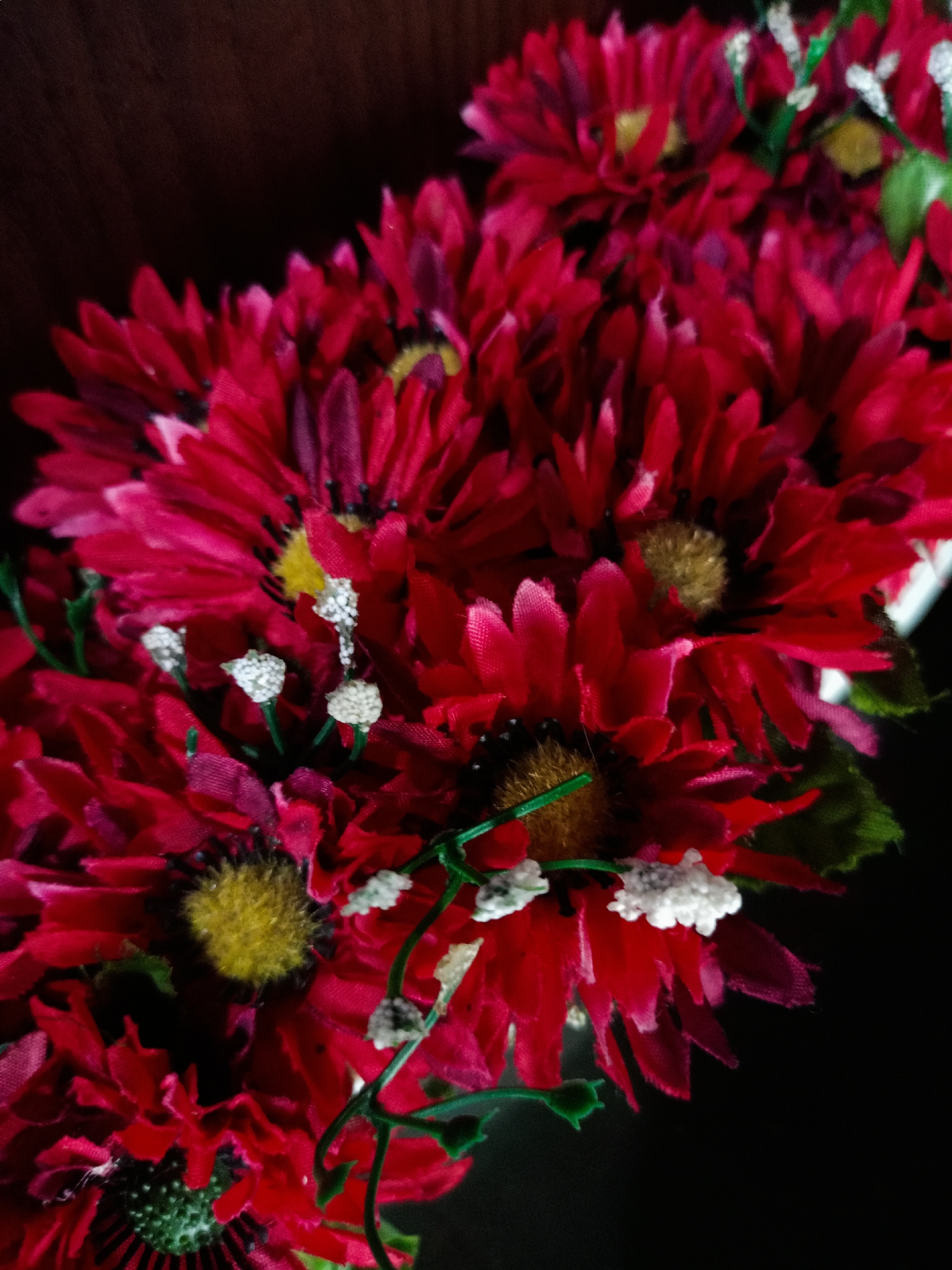 The image shows a collection of red artificial flowers with yellow and brown centers. Some green stems and small white decorative elements are visible among the flowers. The background is dark, appearing to be either a dark surface or a space obscured by shadow. The location and specific details beyond the described objects cannot be determined from the image.