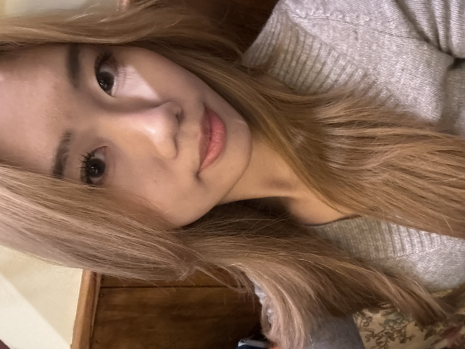 A young East Asian female is depicted in a close-up, chest-up shot within an indoor setting. She has long, straight light brown hair with bangs framing her face, and is looking directly at the viewer with a neutral expression. Her eyes are dark, and her lips are closed with a subtle pink tint. She is wearing a light grey V-neck knitted top with a ribbed texture. The background on the left side features wooden paneling, and a small portion of a muted, patterned fabric, possibly upholstery, is visible behind her left shoulder. The lighting is warm and diffused. The scene is situated in Yuzhong District, China.