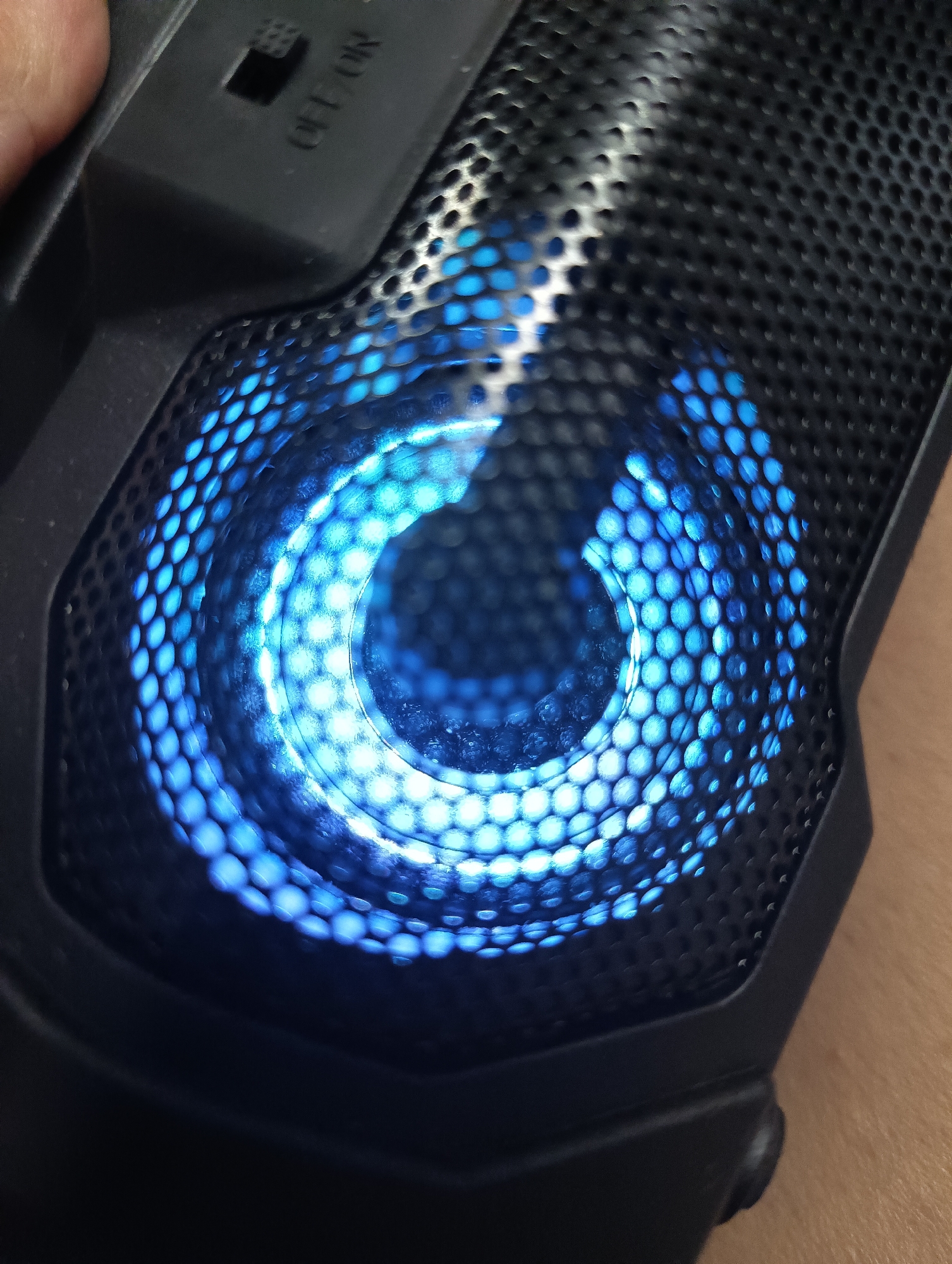 A close-up, slightly angled shot captures an electronic device, likely a portable speaker, with a striking blue illumination. The primary subject is a black, rectangular object featuring a honeycomb-patterned grille over a significant portion of its surface. Through this perforated mesh, a bright, concentric blue light emanates, creating a circular, almost spiral-like glow that is most intense at its center and diffuses outwards.
In the upper left section of the image, a human finger or thumb is partially visible, seemingly holding or interacting with the device. Adjacent to this, above the glowing grille, a black toggle switch with the legible text "OFF ON" is present. The switch appears to be in the "ON" position, indicated by the visible lettering and the active light. The device itself is a dark, matte grey or black, with a slightly textured finish.
The setting appears to be indoors, with a light brown surface, possibly skin or wood, visible in the bottom right corner, suggesting the device is being held or placed on a surface. The vibrant blue light is the focal point, indicating the device is powered on and active. No specific time of day or weather conditions are discernible from the image content itself, beyond the internal illumination. The location context is Biliaivka, Ukraine.
