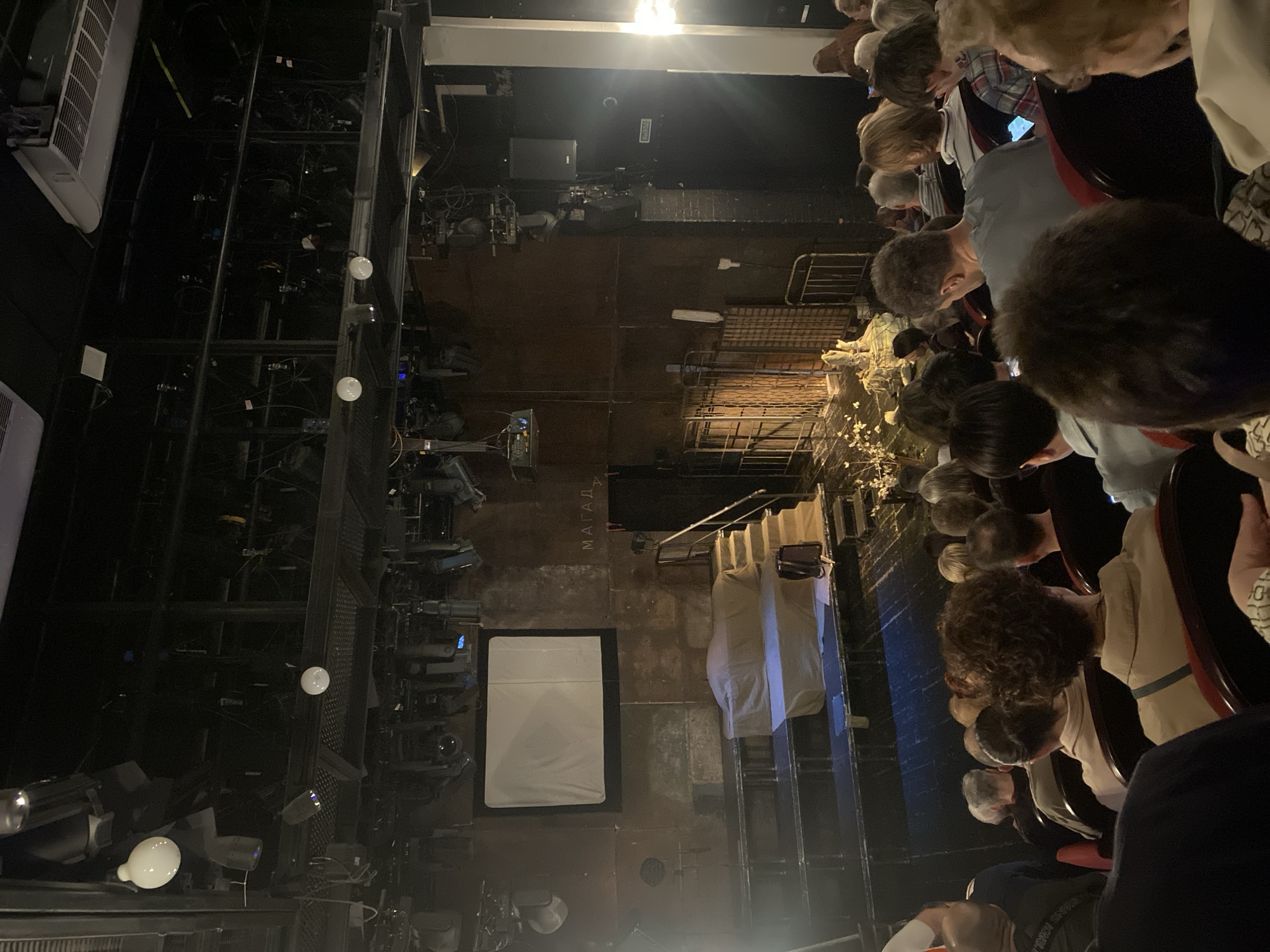 From an elevated perspective, the image captures the interior of the Theatre of Nations in Moscow, Russia. A section of audience members are visible in the right foreground, heads tilted down, some holding phones. A stage is set up with props, including white fabrics and metal frames, illuminated by stage lighting. Technical equipment such as light rigging and theatrical lights are visible to the left. A projection screen with a white display is present. "MAYAJA" is written on the floor.