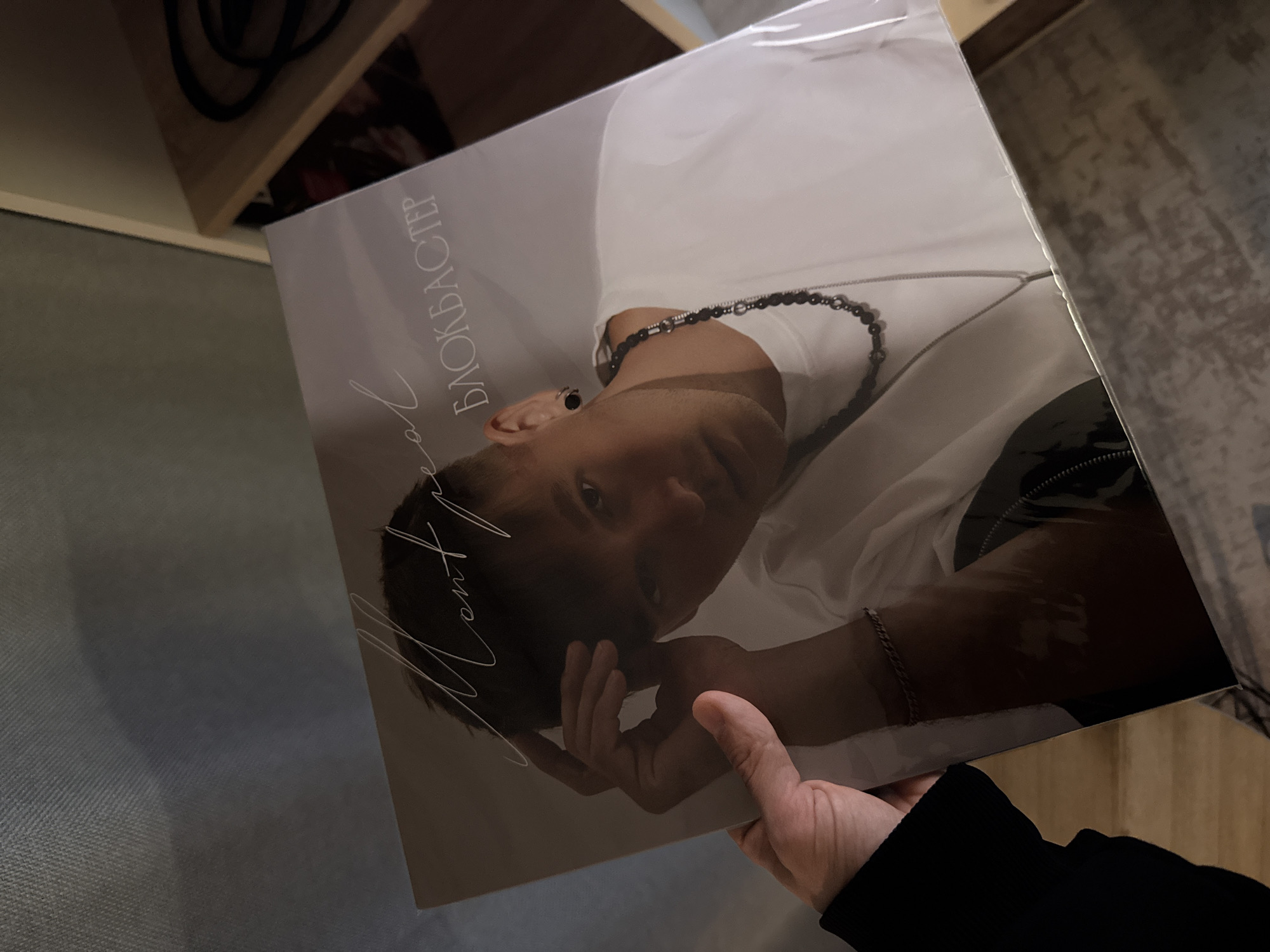 A photograph taken indoors shows a person holding a vinyl record album. The album cover features a man with short, dark hair wearing a white shirt and a black beaded necklace. The title "MONTREAL" is written vertically along the left side of the cover, followed by the word "БЛОКБАСТЕР" written vertically in Cyrillic. The album is partially covered in plastic wrap. The person holding the album is wearing a black long-sleeved shirt. The background includes a wooden shelf with visible objects and a wooden surface. The location is not specified in the image or its context.
