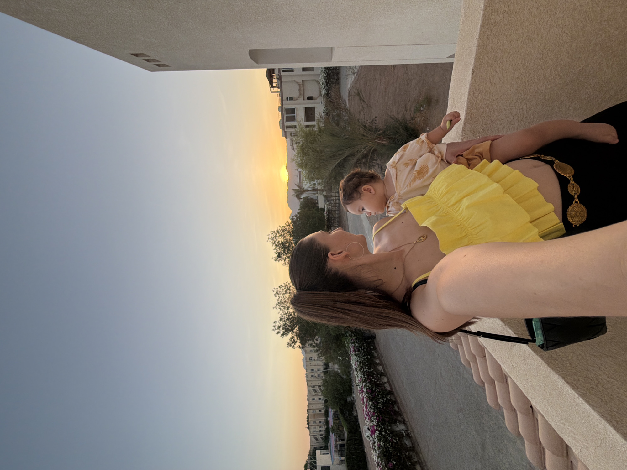 A woman is holding a baby on a balcony in Sharm El Sheikh, Egypt, during sunset. The woman has her hair pulled back in a high ponytail and is wearing a yellow ruffled top, a gold chain necklace, and a gold coin belt. The baby is wearing a light-colored patterned shirt and shorts. In the background, there are buildings, palm trees, and mountains in the distance. The sky is a gradient of blue and orange, indicating the sun is setting.