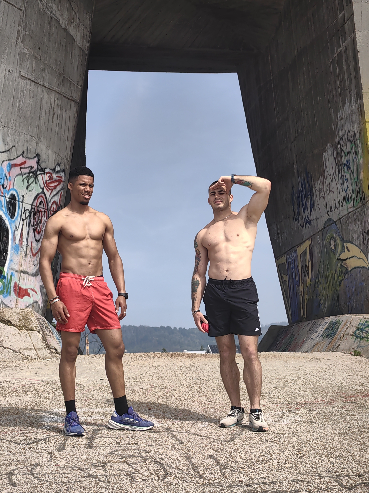 The image displays two shirtless adult males standing on a gravel-covered surface beneath the concrete archway of the Asparuhov Bridge in Varna, Bulgaria. The imposing concrete pillars of the bridge flank the scene, both heavily marked with vibrant, multi-colored graffiti.
The male on the left, positioned closer to the viewer, has short dark hair, a muscular physique, and wears red shorts with white drawstrings, black ankle socks, and blue and purple athletic shoes. He sports a black wristwatch on his left wrist and multiple bracelets on his right wrist, along with a ring. He looks towards the right side of the frame with a subtle smile.
The male on the right, positioned slightly further back and to the right, also has short dark hair and a muscular build, with visible tattoos on both arms. He wears black athletic shorts featuring a white Nike swoosh, and light-colored athletic shoes with black laces. He has a chain around his neck, a dark wristwatch on his left wrist, and a ring on his left hand. His left hand is raised, shielding his eyes, and he holds a small red object in his right hand.
The background, visible through the bridge's arch, shows a clear sky transitioning to a hazy horizon with distant hills. The scene is illuminated by natural daylight, casting faint shadows on the ground.