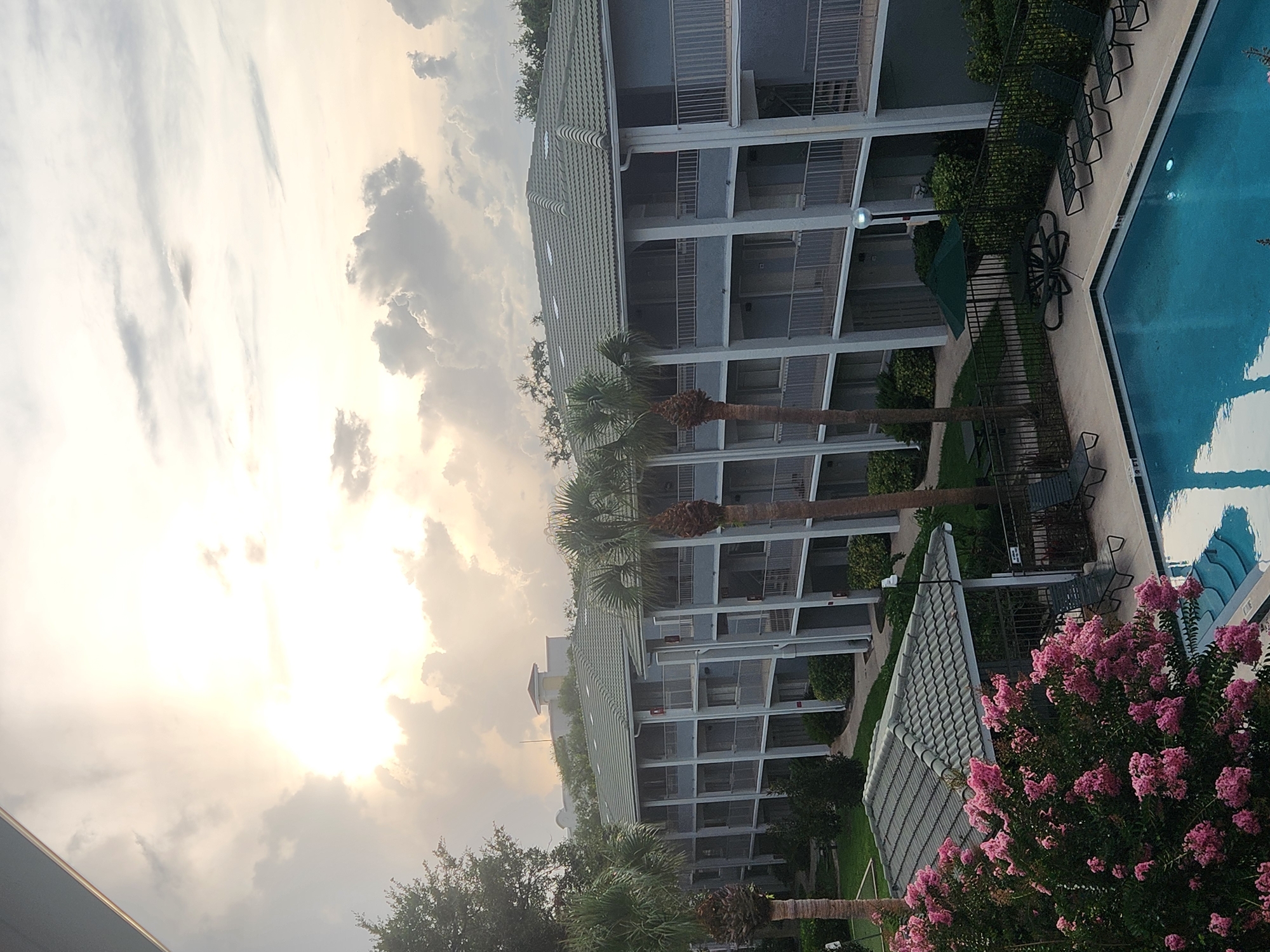 The image depicts a view from an elevated vantage point overlooking a courtyard area of a hotel or resort in Altamonte Springs, Florida, USA. The foreground features a vibrant pink flowering bush and a portion of a bright blue swimming pool. Beyond the pool, a low-rise building with multiple stories and balconies is visible. The building has a light gray exterior and a green tiled roof. Several palm trees are interspersed around the building and in the courtyard, with some appearing mature and tall.
The sky above is dramatic, filled with heavy, textured clouds illuminated by what appears to be the sun breaking through or setting. The lighting suggests it could be late afternoon or early morning, with a diffused, soft glow. In the background, beyond the main building, other structures and trees are visible, hinting at a developed area. The scene is tranquil, with no people or active events captured. The overall impression is a resort or hotel setting during a period of changing weather.