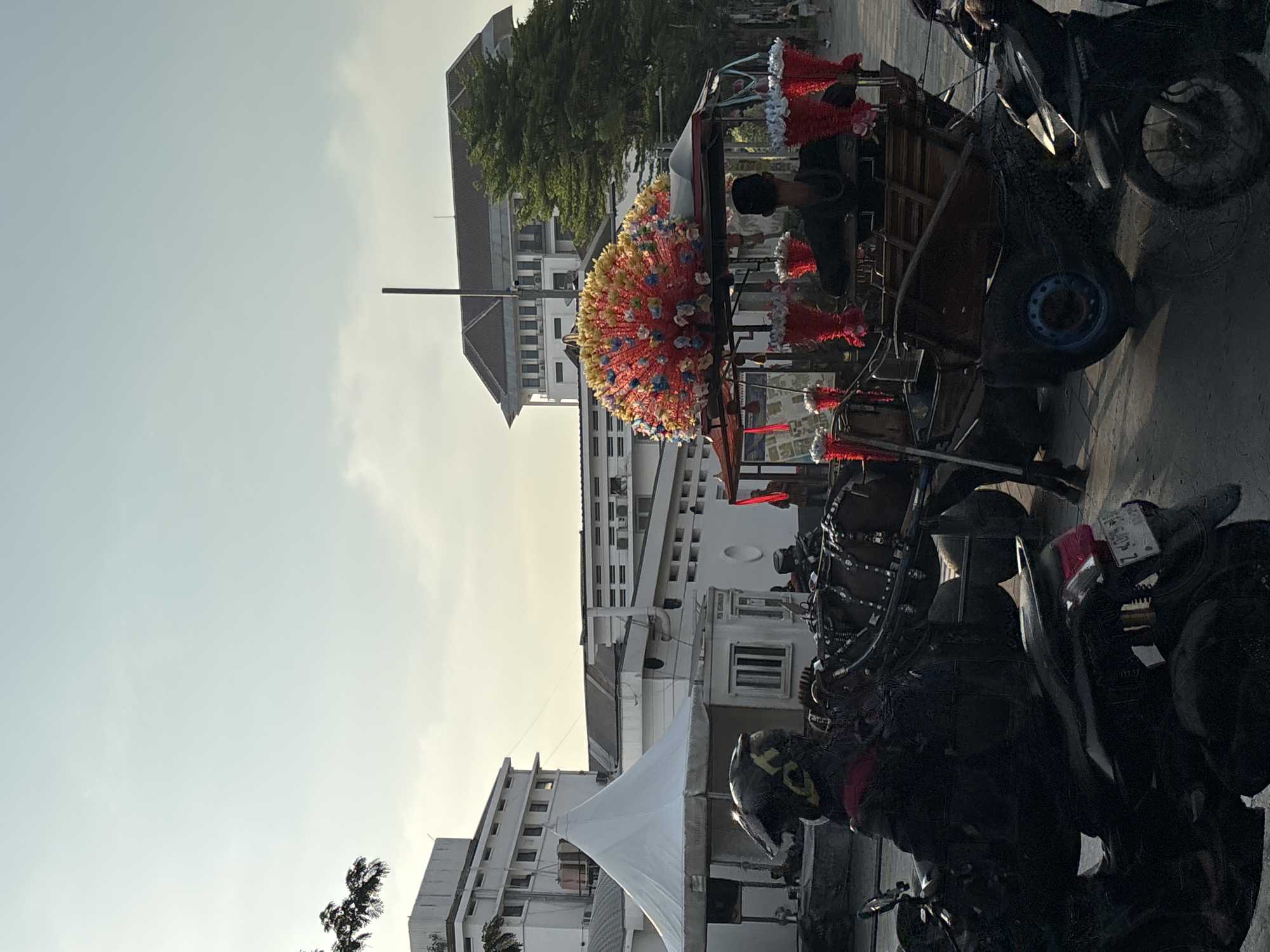 This is a medium-shot, eye-level, vertical, natural light photograph taken during the day, possibly late afternoon, in the Kota Tua (Old Town) area of West Jakarta, Indonesia. The scene features a horse-drawn carriage decorated with a large, vibrant, fan-shaped arrangement of red, yellow, and blue flowers on its roof. The carriage itself is made of dark wood and has a red awning. Several red floral bouquets are attached to the sides of the carriage.
In the foreground, a person wearing a black helmet with yellow accents is seated on a black motorcycle. The motorcycle's license plate is partially visible and appears to have the characters "Z 4075 M". Another black motorcycle is parked on the right side of the frame.
Behind the carriage, two people are visible, one appearing to be the carriage driver wearing a dark cap. In the background, several white buildings with multiple windows and air conditioning units are prominent. One building has a large white tent structure in front of it. A tall, thin metal pole stands behind the carriage, extending towards the sky. Trees with green foliage are visible on the right side of the image. The sky is a pale blue with some hazy clouds. The overall impression is a street scene in a historic urban setting with a touch of traditional transport.