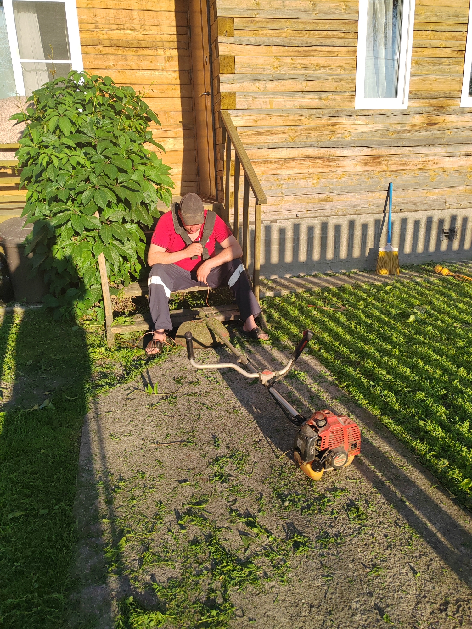 The image depicts a man sitting on a wooden bench outside a wooden house. He is wearing a red shirt, dark pants with a white stripe, a cap, and sandals. He is looking down and appears to be resting. In front of him, on the ground, is a red string trimmer. The ground is covered with cut grass, indicating recent yard work. A large green bush is to the man's left. To the right of the house, a yellow broom with a blue handle stands upright against the wall. The house has wooden siding and white-framed windows. The sun is casting long shadows on the ground, suggesting it is late afternoon or early morning. The overall scene appears to be a residential outdoor setting. There is no visible text in the image.