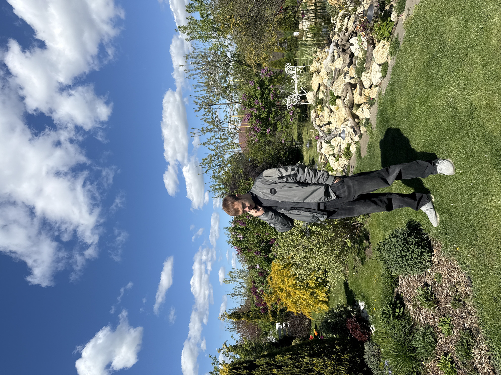 A young man stands outdoors in a garden in Pervorechenskoye, Russia, under a bright blue sky with scattered white clouds. The image is taken on a sunny day, evidenced by the clear light and distinct shadows cast on the grass.
The man, who appears to be in his late teens or early twenties, is positioned slightly off-center to the left, facing towards the right side of the frame. He is wearing a dark grey or black jacket over dark pants and light-colored sneakers. His right hand is raised to his face, near his mouth or chin, in what appears to be a pensive or observant gesture.
The background features a well-maintained garden with a vibrant mix of green lawn, various trees, and bushes with different foliage colors, including bright yellow and deep green. There are purple flowering plants, possibly lilac, and other diverse greenery. To the right, there is a prominent rockery with light-colored stones, some driftwood, and small decorative garden lights interspersed among the rocks. A white, ornate metal garden bench is visible near the center-right, tucked among the plants and rocks. The far background shows glimpses of what appears to be a residential roof and more distant trees, suggesting a suburban or rural setting. There is no visible text in the image.