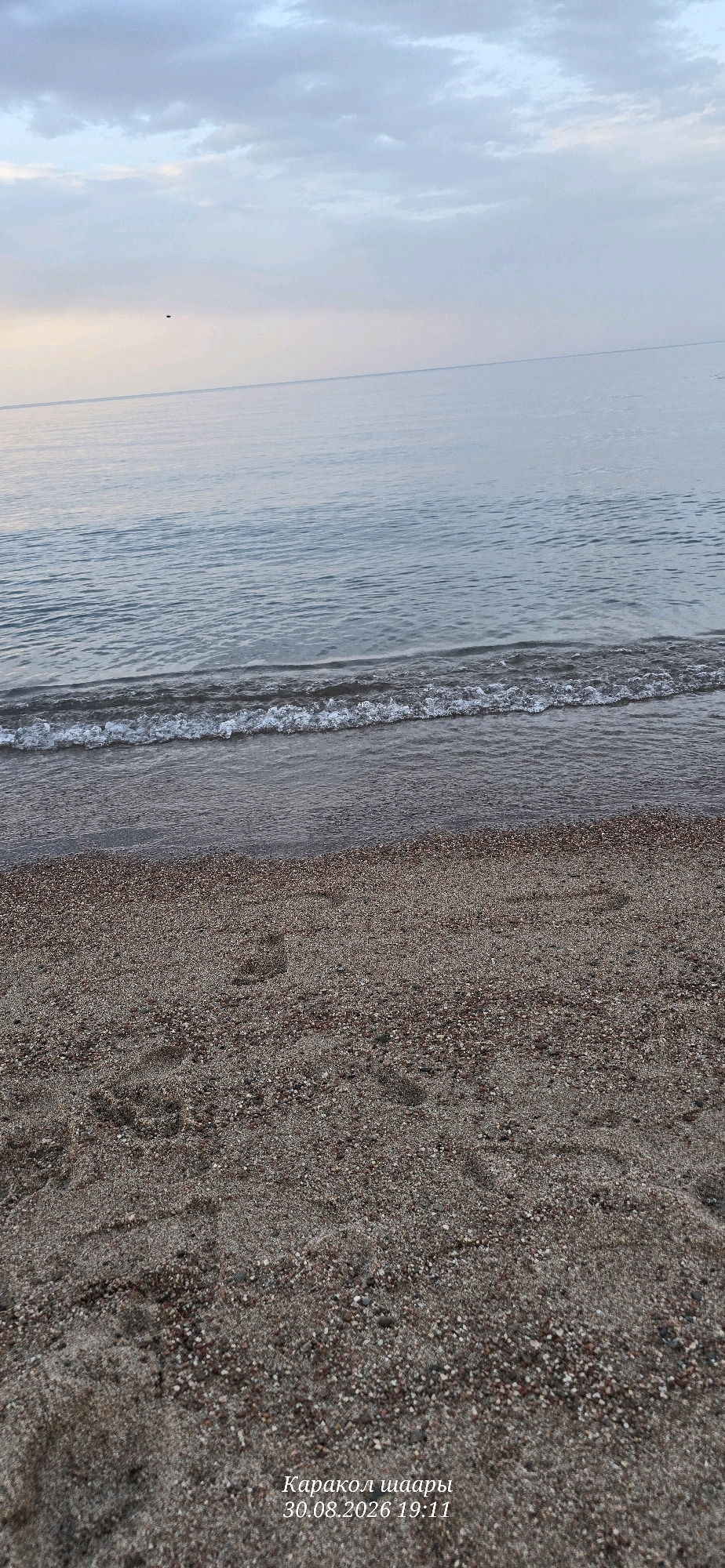 The image depicts a tranquil scene at a beach, likely in the city of Тосор, Kyrgyzstan, as indicated by the text overlay. The foreground is dominated by a sandy and pebbled beach with visible footprints, suggesting recent human presence. A gentle wave is lapping at the shore, creating a foamy white line where it meets the land. The water transitions into a calm, expansive body of water that stretches to the horizon. The sky above is overcast with soft, diffused clouds, creating a serene and somewhat muted atmosphere. The overall impression is one of peace and natural beauty. The text overlay "Каракол шаары 30.08.2026 19:11" indicates the location as Karakol city and the date and time as August 30, 2026, at 7:11 PM.