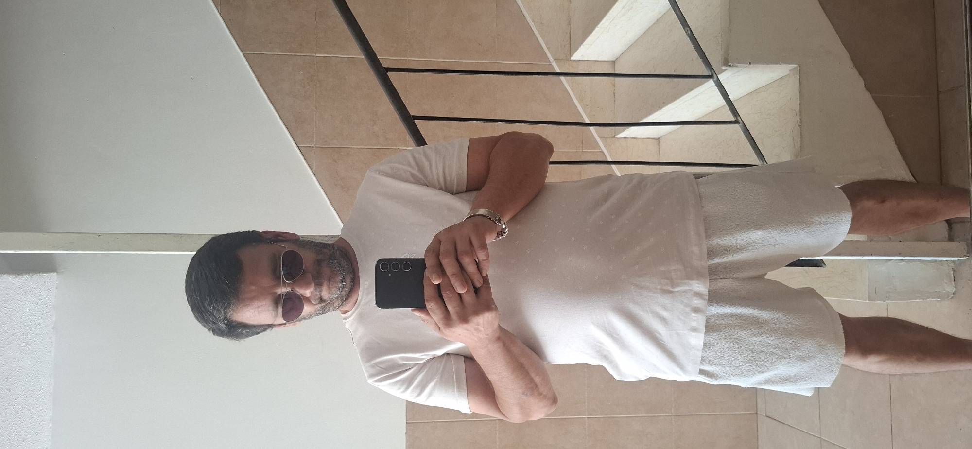 A man is taking a mirror selfie. He is wearing a light-colored t-shirt and white shorts. He has sunglasses on and is holding a smartphone in his hands. He is standing in front of a tiled wall with a staircase in the background. The environment appears to be indoors, possibly a home or a public building. Based on the tiled walls and the style of the staircase, it's plausible this is in Yerocham, Israel.