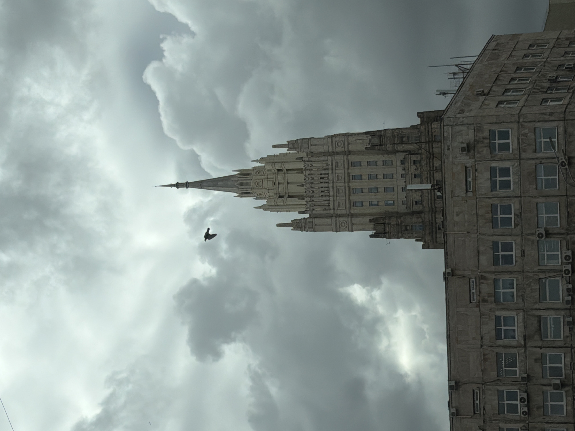 This image, taken in Moscow, Russia, showcases a towering Stalinist-era skyscraper under a dramatically overcast sky. The photograph is oriented vertically, emphasizing the upward reach of the building against the dense cloud cover.
The primary subject is a majestic, ornate skyscraper, a distinctive example of Moscow's "Seven Sisters" architectural style. Its facade, appearing to be stone or concrete, is richly detailed with multiple tiers and regularly spaced windows. A prominent, multi-faceted spire crowns the building, tapering to a sharp point that extends high into the clouds. Adjacent to and slightly to the right of this grand structure, another multi-story building stands. It shares a similar aged appearance, featuring numerous rectangular windows, many of which have external air conditioning units mounted beneath them.
The sky is a dominant element, filled with heavy, turbulent grey clouds that suggest either an impending storm or a generally gloomy, cloudy day. Despite the extensive cloud cover, some lighter patches are visible, indicating diffused daylight. A single bird, silhouetted in dark contrast against the brighter areas of the clouds, is captured in flight to the left of the main building's spire.
The scene depicts an urban environment, likely a historic part of Moscow, characterized by its imposing architecture and somber weather. No people or visible text are discernible in the image. The aged appearance of the buildings contributes to a sense of historical gravitas under the diffused light of the cloudy day.