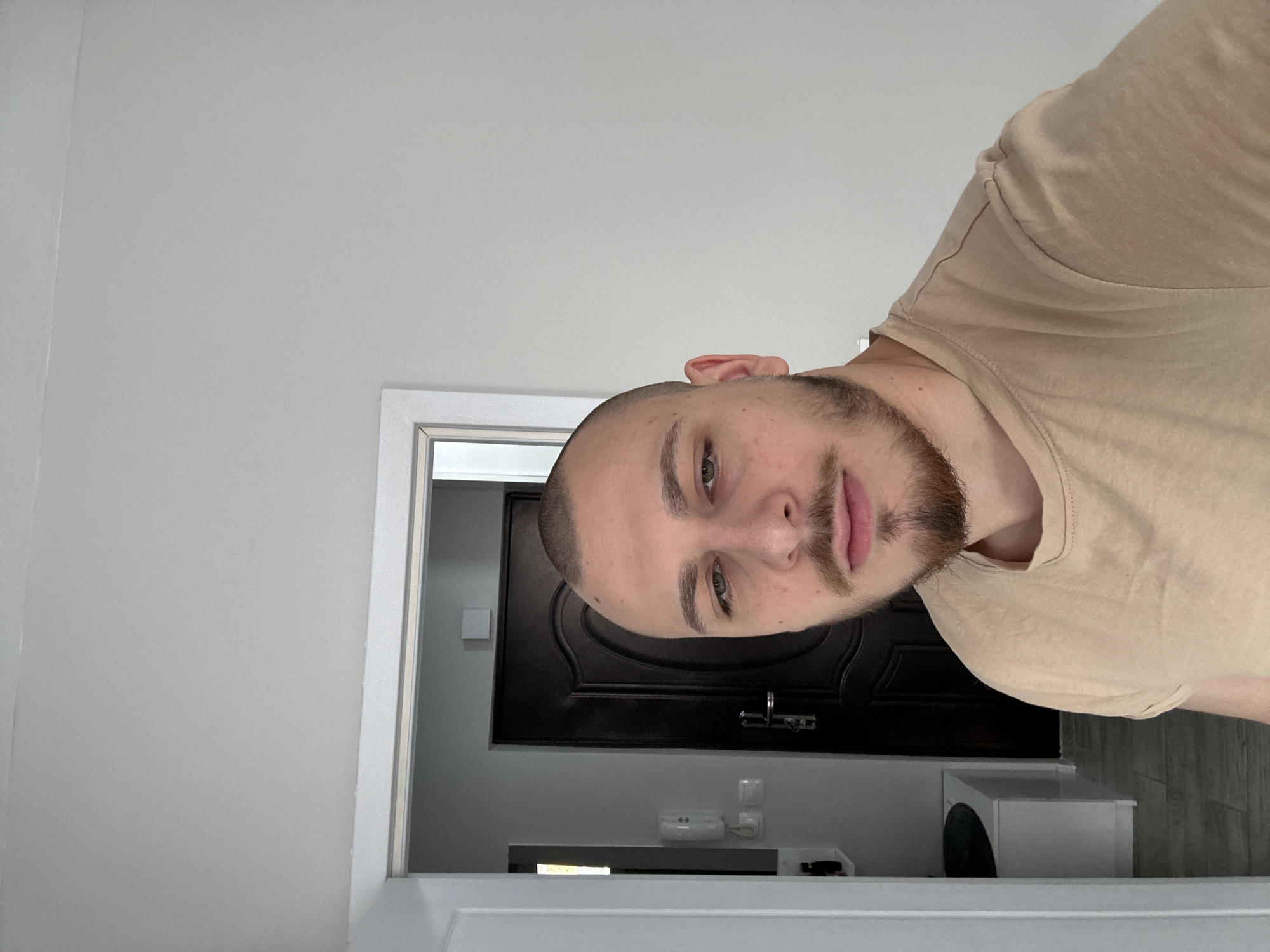 An indoor scene in Stara Zagora, Bulgaria, depicts a male individual in a head-and-torso shot, facing the camera directly. The subject possesses a closely shaved head and a dark goatee-style beard connected to a mustache, with light-colored eyes. He wears a plain, light tan, short-sleeved t-shirt. In the background, to the left of the subject, an open doorway reveals a dark brown, paneled interior door with a silver handle and lock mechanism. Above this door, a light-colored square object is mounted on the wall. To the left of the dark door, a white wall-mounted intercom unit is visible, with a smaller white switch-like object positioned below it. Further to the left, through an additional opening, a partial view of an adjacent room shows a white utility cabinet with a dark circular top and light-toned floor tiles. The walls surrounding the subject and in the visible background are painted white.
