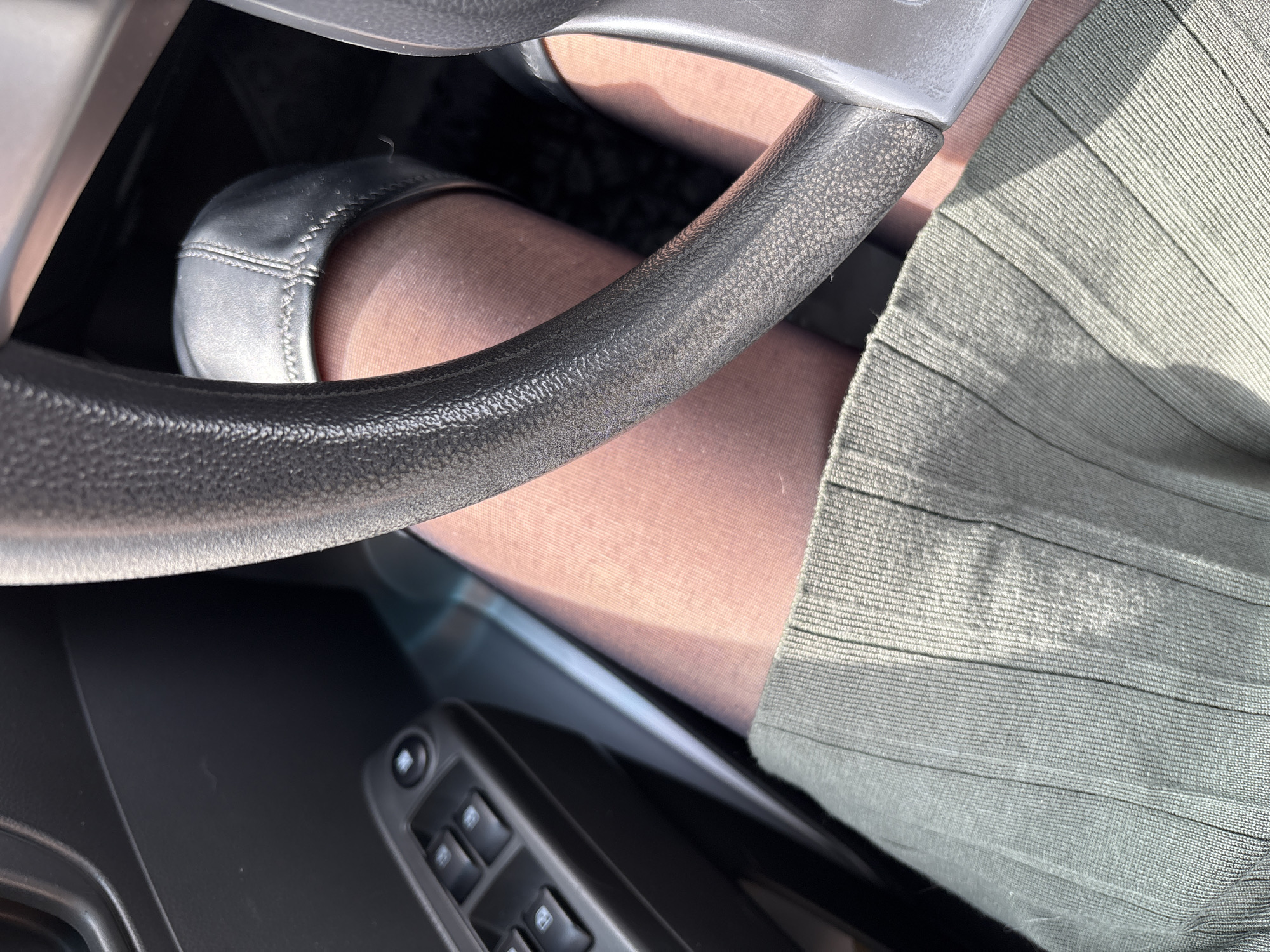 The image shows a view from inside a car, focusing on the driver's side. The steering wheel is prominently displayed in the foreground. A leg covered in sheer pantyhose is visible, positioned under the steering wheel, with a black flat shoe on the foot. Part of a ribbed, possibly green, skirt is also visible. A portion of the car's interior, including the window control panel, is in the lower left corner of the image. The location is not discernible from the visual information provided.