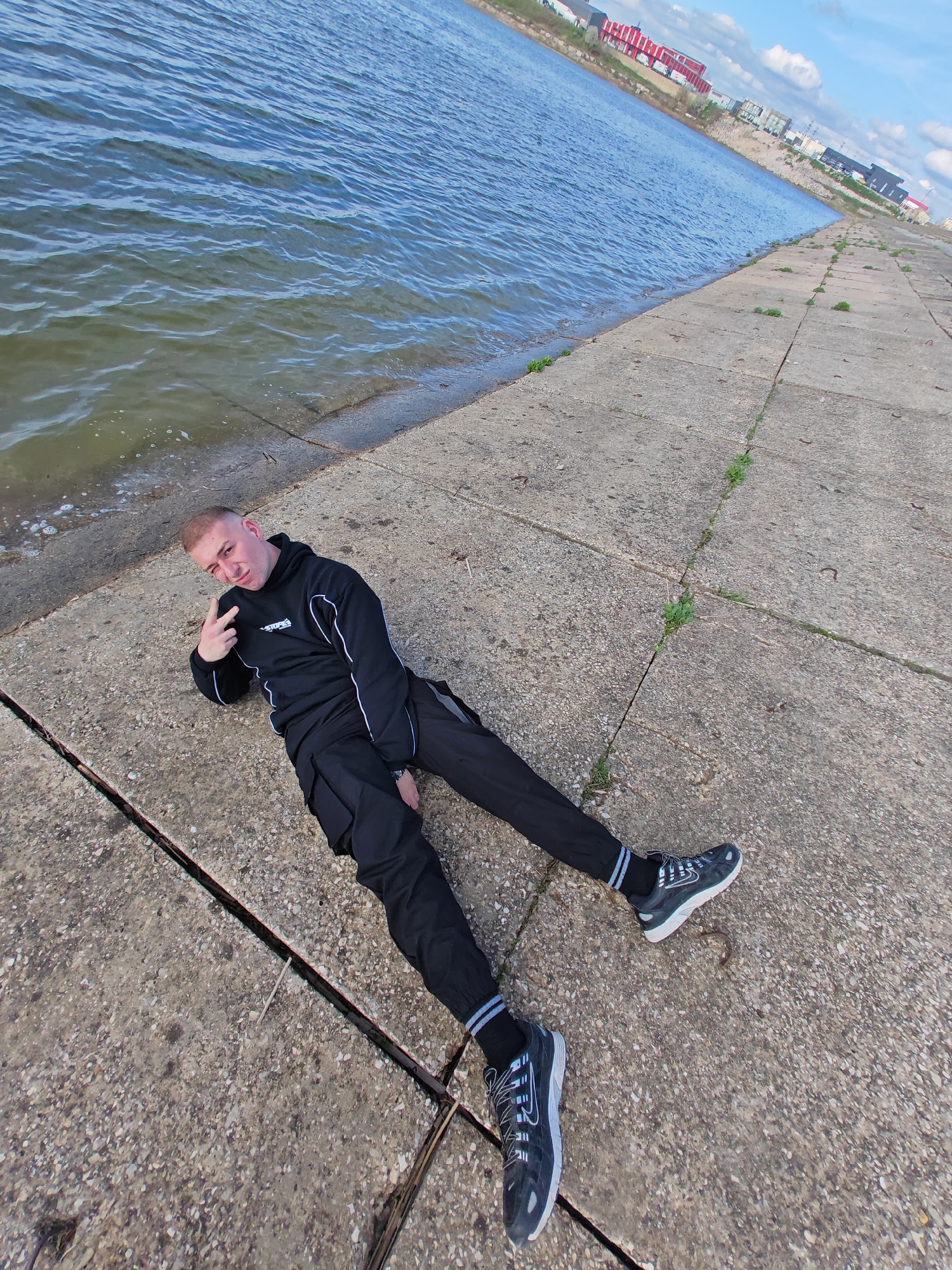 A young man with a shaved head is lying on a concrete pier or quay, adjacent to a body of water. He is wearing a black hooded sweatshirt with white trim, black pants, and black athletic shoes with light-colored accents. He is facing the camera and holding up two fingers in a peace sign. The pier is constructed of rectangular concrete blocks with cracks and occasional small plants growing in the gaps. In the background, across the water, are buildings of various colors, primarily red and white. The sky is partly cloudy. The location appears to be in an urban area. The city of Shumen, Bulgaria, is not visually identifiable in the image.